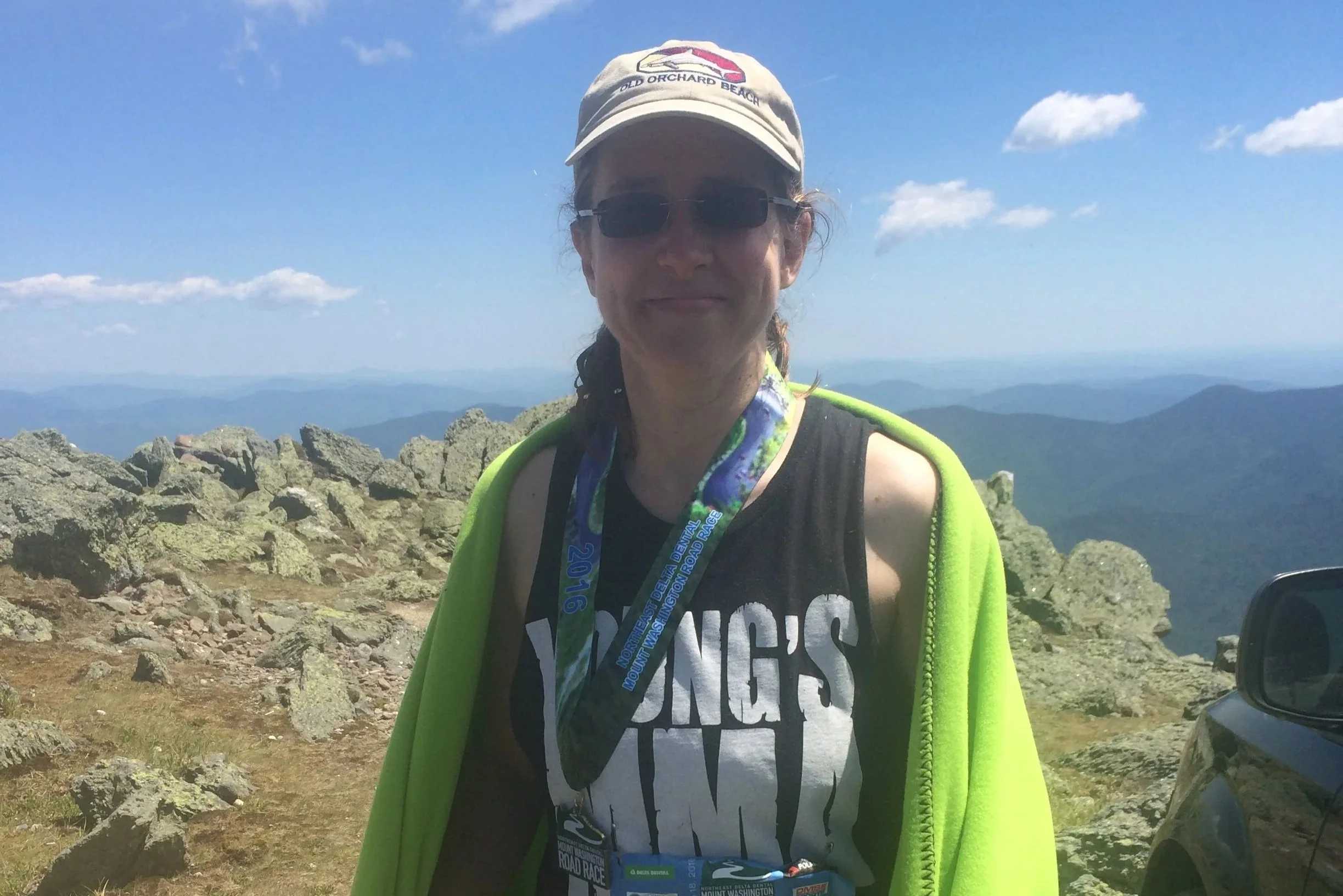A woman wearing sunglasses, a cap with 'Old Orchard Beach' logo, and a black tank top with white text stands outdoors on a mountain ridge with rocks and a scenic distant view of mountains under a partly cloudy sky.