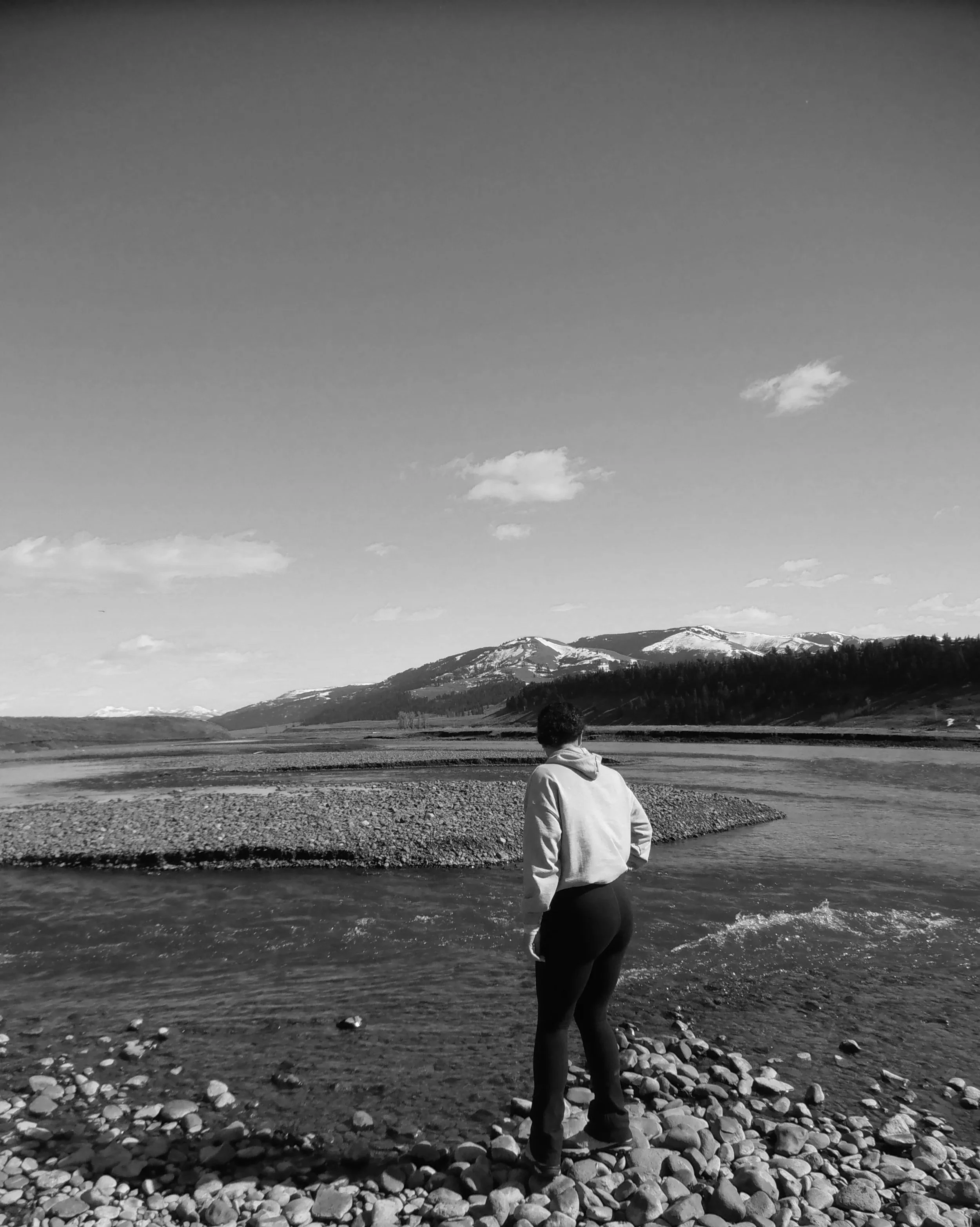 A person standing by a river with mountains in the background, in black and white.
