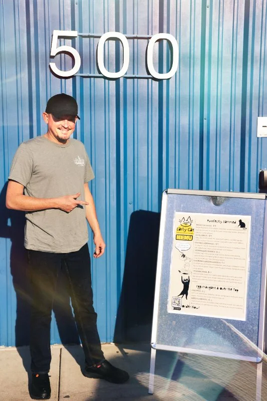 A man smiling and pointing at a signboard outside a building with a blue metal wall. The number 500 is displayed above him.