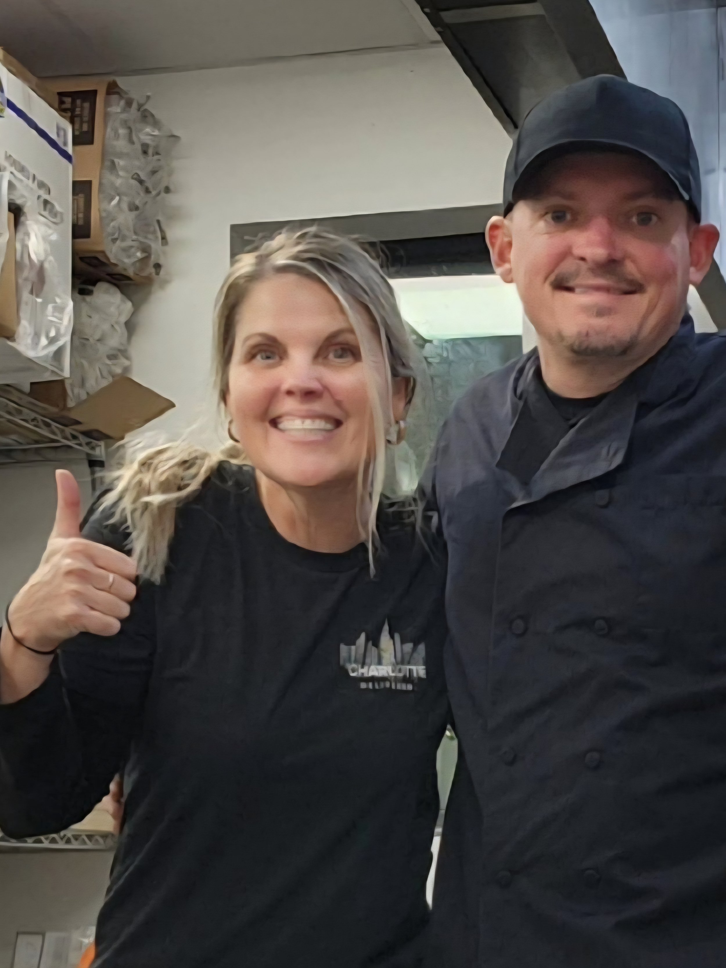 Woman and man in a kitchen, smiling and taking a selfie. The woman is giving a thumbs-up. The woman is wearing a black shirt with a logo that says 'Charlotte' and the man is in a black chef's jacket and cap.