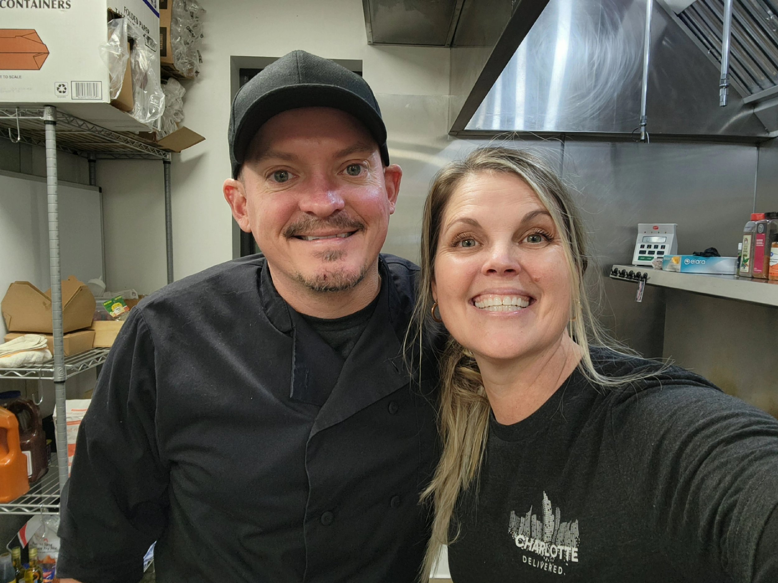 A man and woman smiling, taking a selfie in a commercial kitchen. The man is wearing a black chef's coat and a black cap. The woman is wearing a black shirt with a Charlotte skyline graphic.