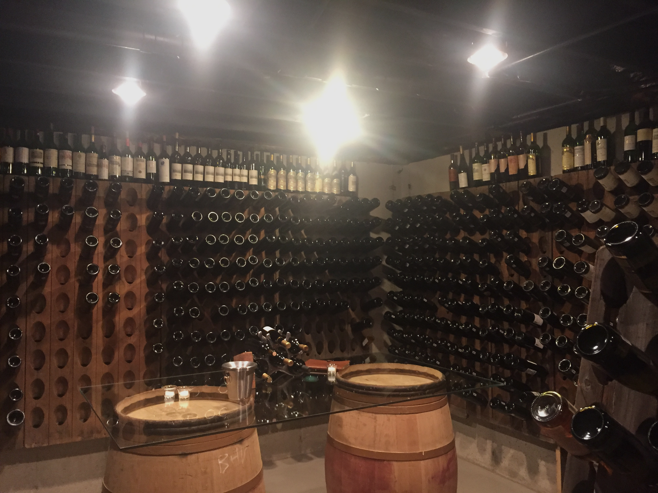 Wine bottles stored on wooden racks in a wine cellar, with a barrel table and a glass tasting setup.