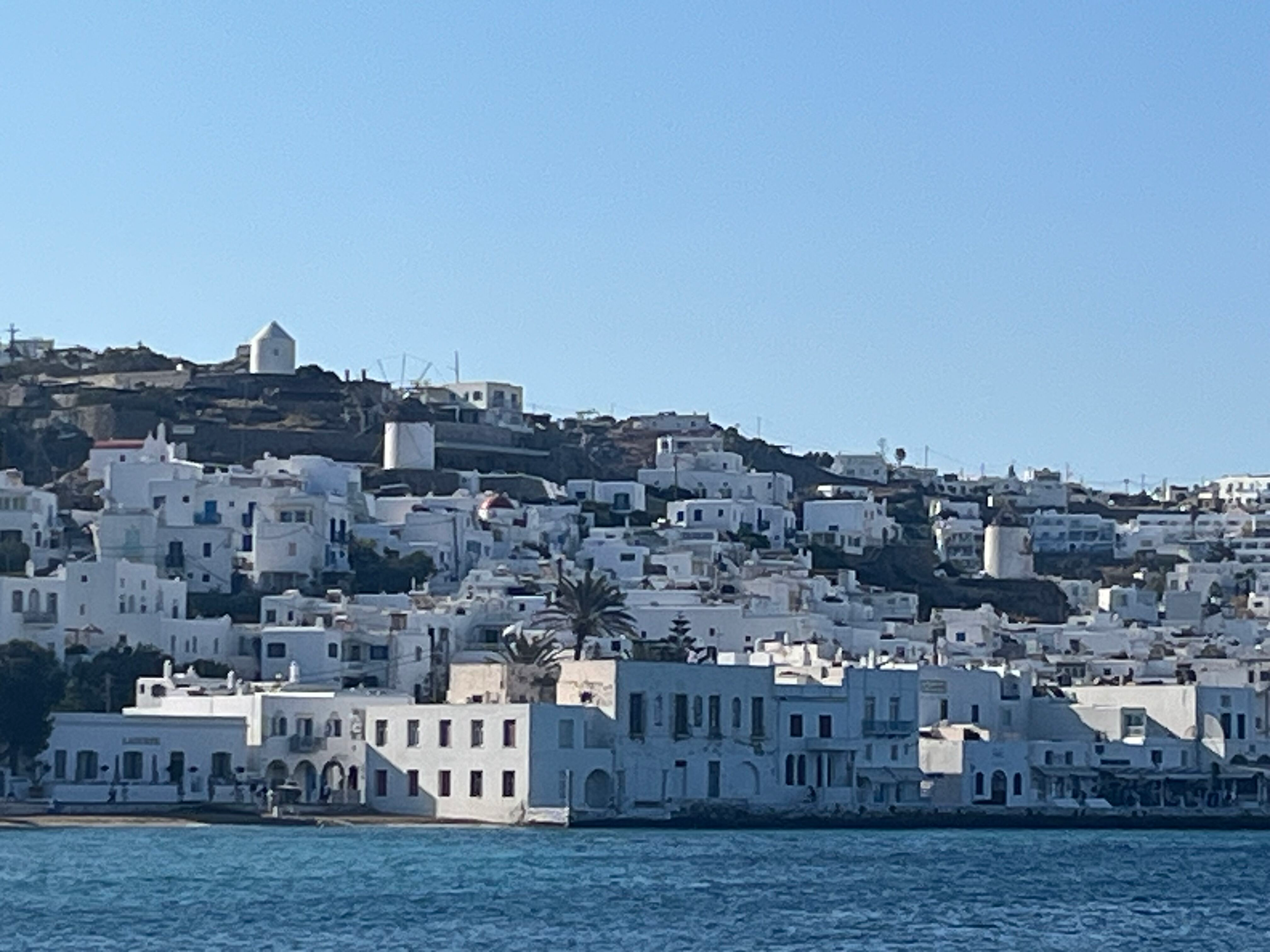 View of a hillside town with white buildings, some with flat roofs, along a waterfront with blue water in the foreground under a clear blue sky.
