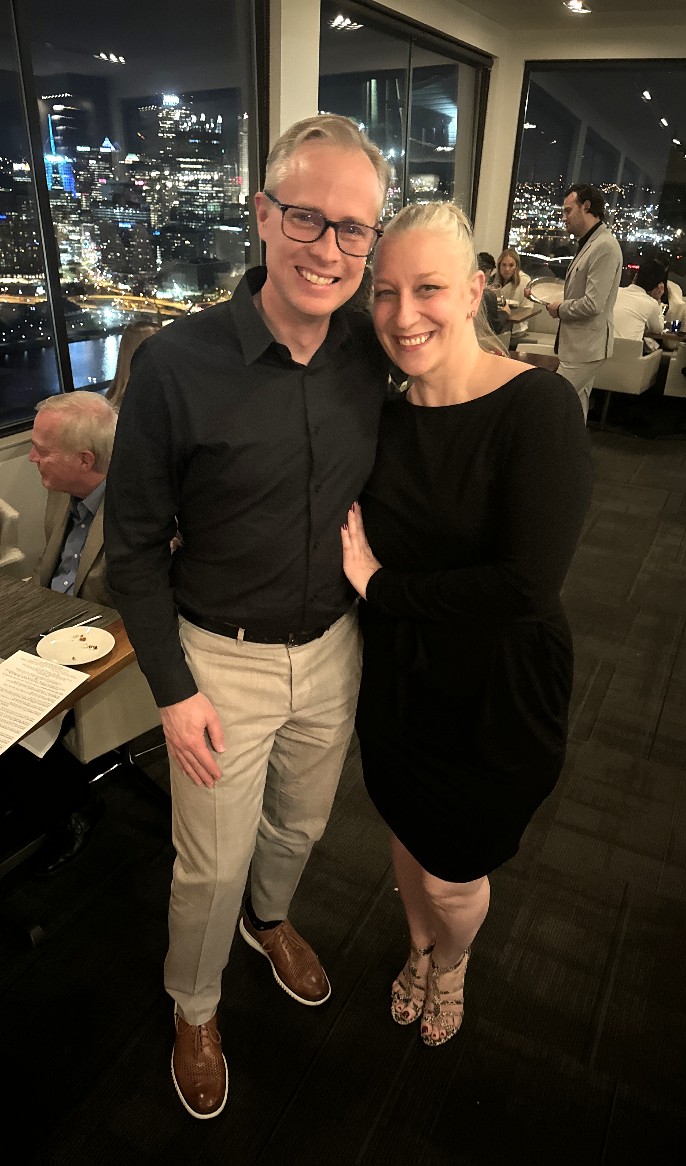A smiling man and woman standing close together at Altuis restaurant at night with Pittsburg skyline view through large windows behind them.