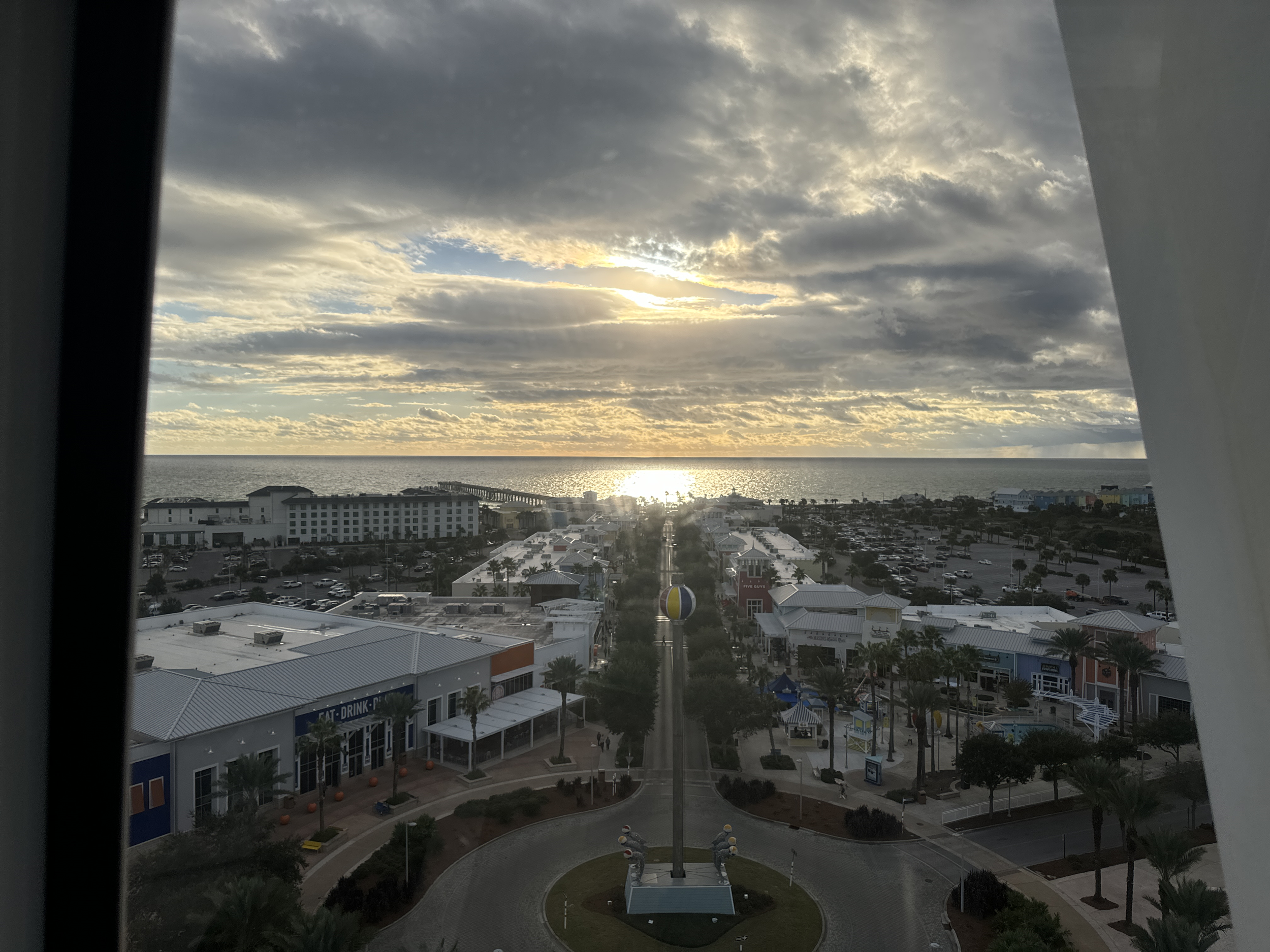 View of a coastal town taken from a high vantage point, with buildings, parked cars, trees, and umbrellas, overlooking the ocean under a cloudy sky during sunset.