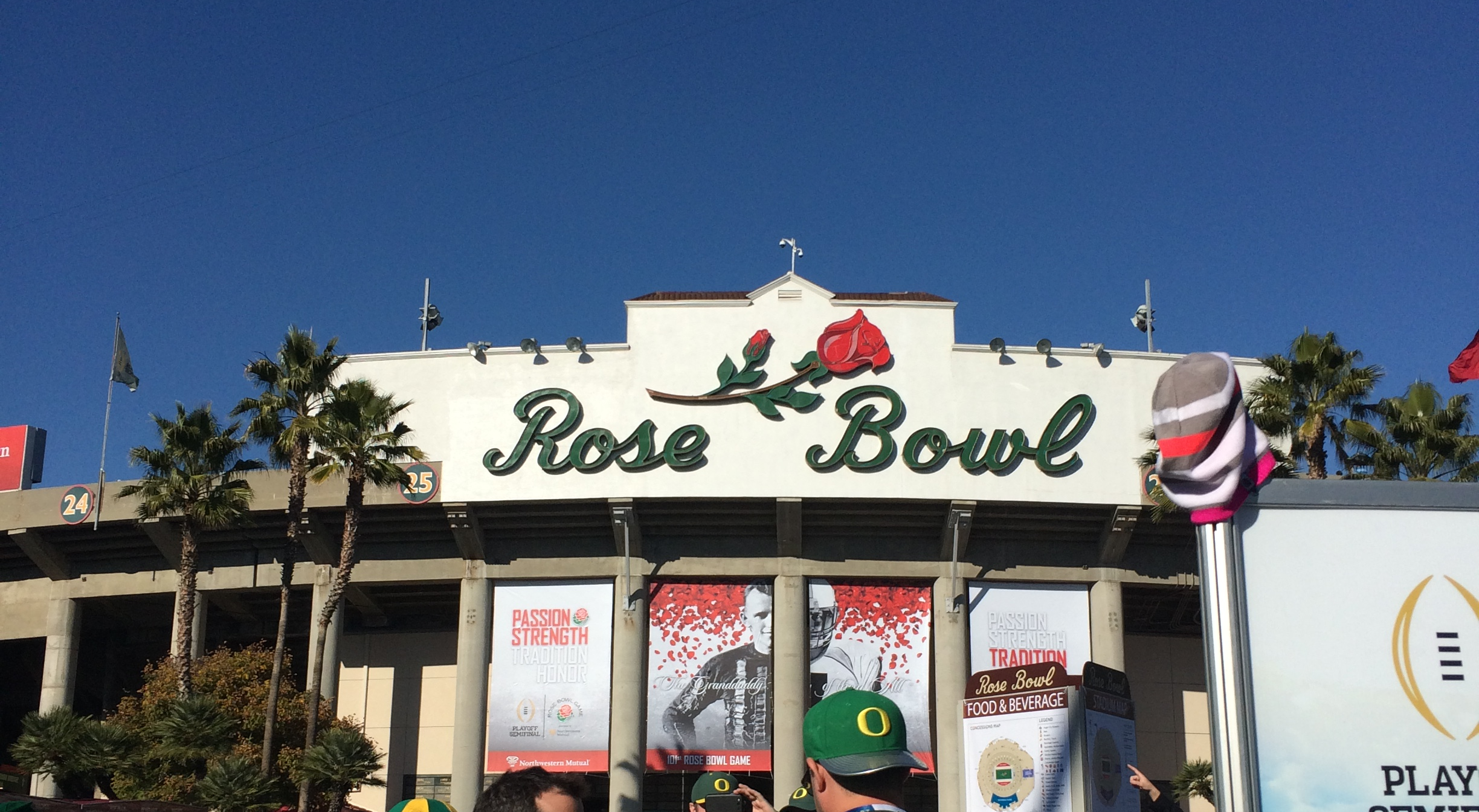 The entrance of Rose Bowl stadium with large green 'Rose Bowl' sign and a red rose logo, palm trees on either side, and a crowd of people wearing green hats in front, with banners advertising football and the Rose Bowl game.