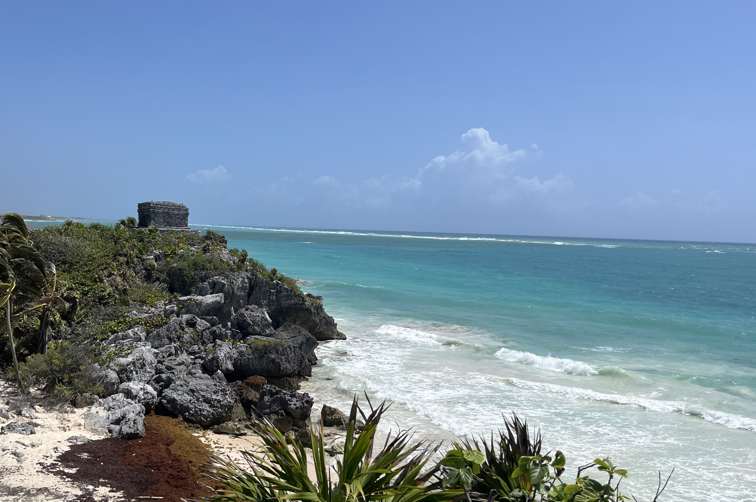 Tropical beach with rocky shoreline, green vegetation, turquoise ocean, and a stone structure on the cliff, under a blue sky with clouds.