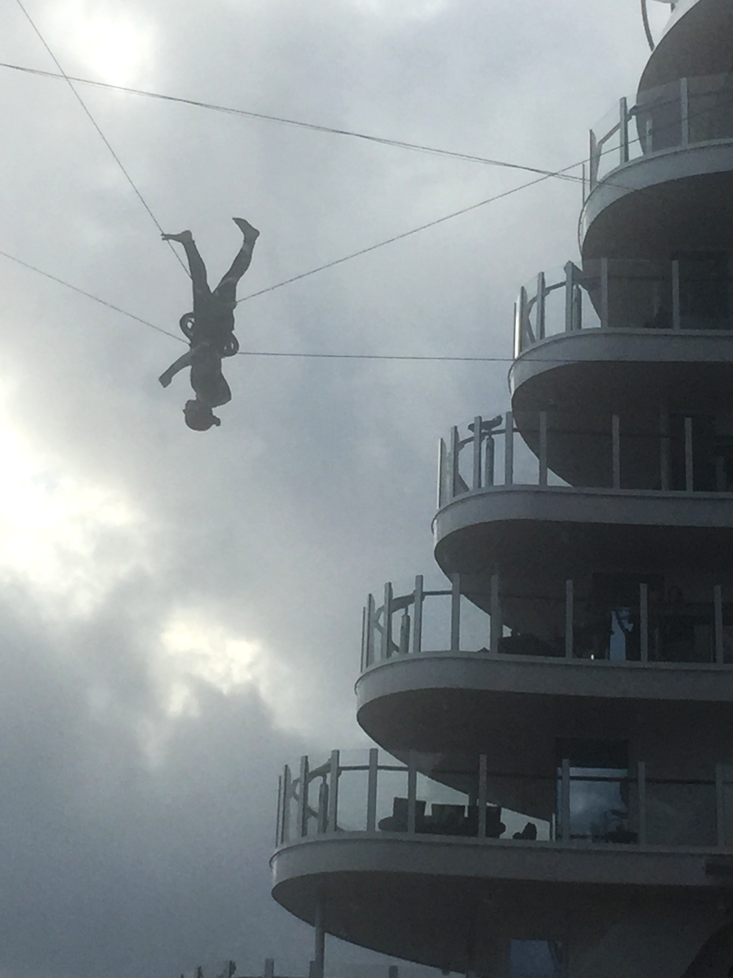 Person zip lining outdoors next to a multi-story cruise ship with balconies, under a cloudy sky.