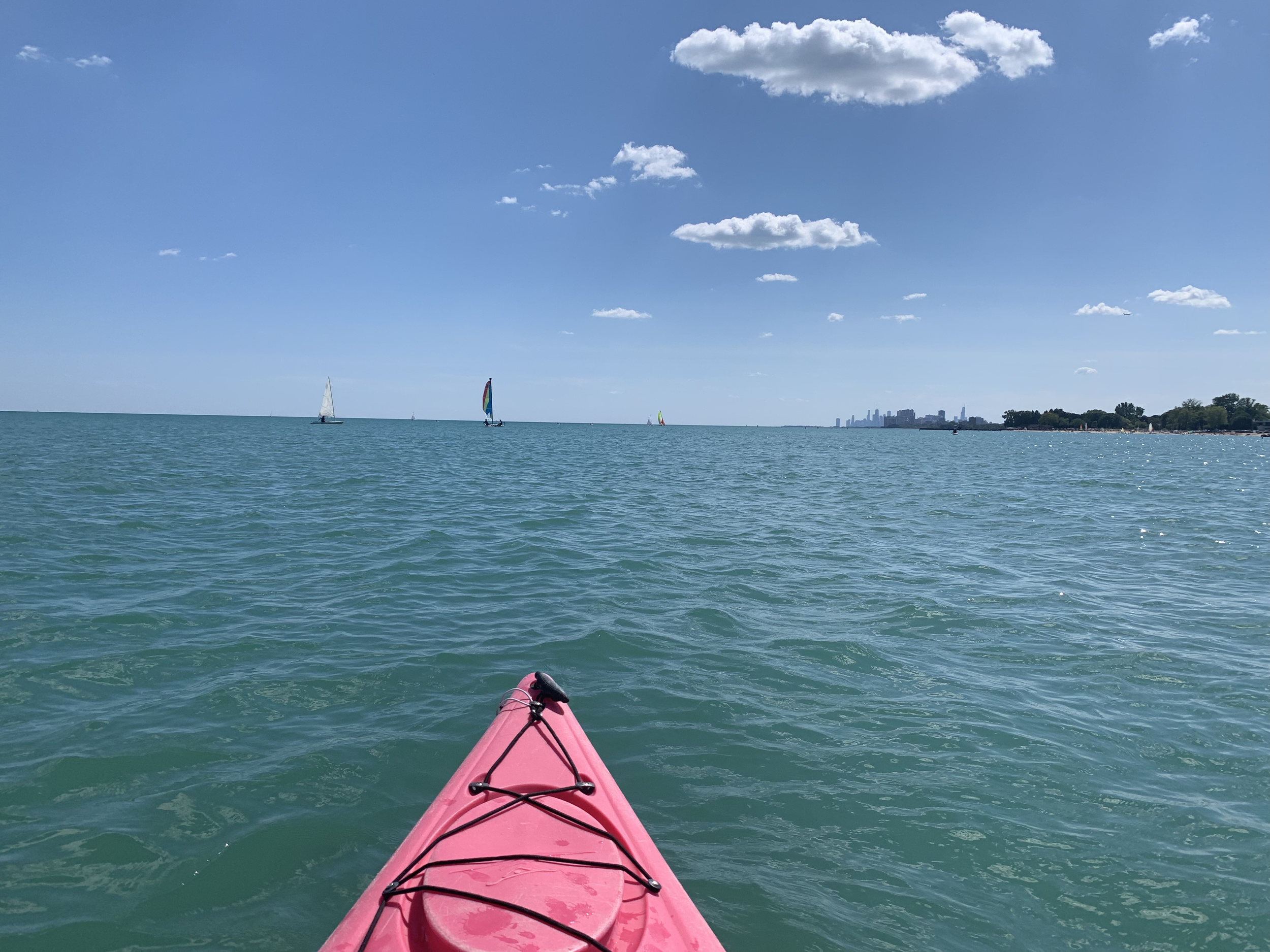 View from a pink kayak on a calm body of water with sailboats in the distance, a partly cloudy blue sky, and Chicago skyline on the horizon.