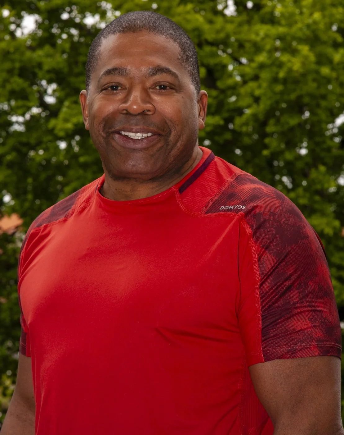 A smiling man with dark skin and short hair, wearing a red athletic shirt, outdoors with green trees in the background.