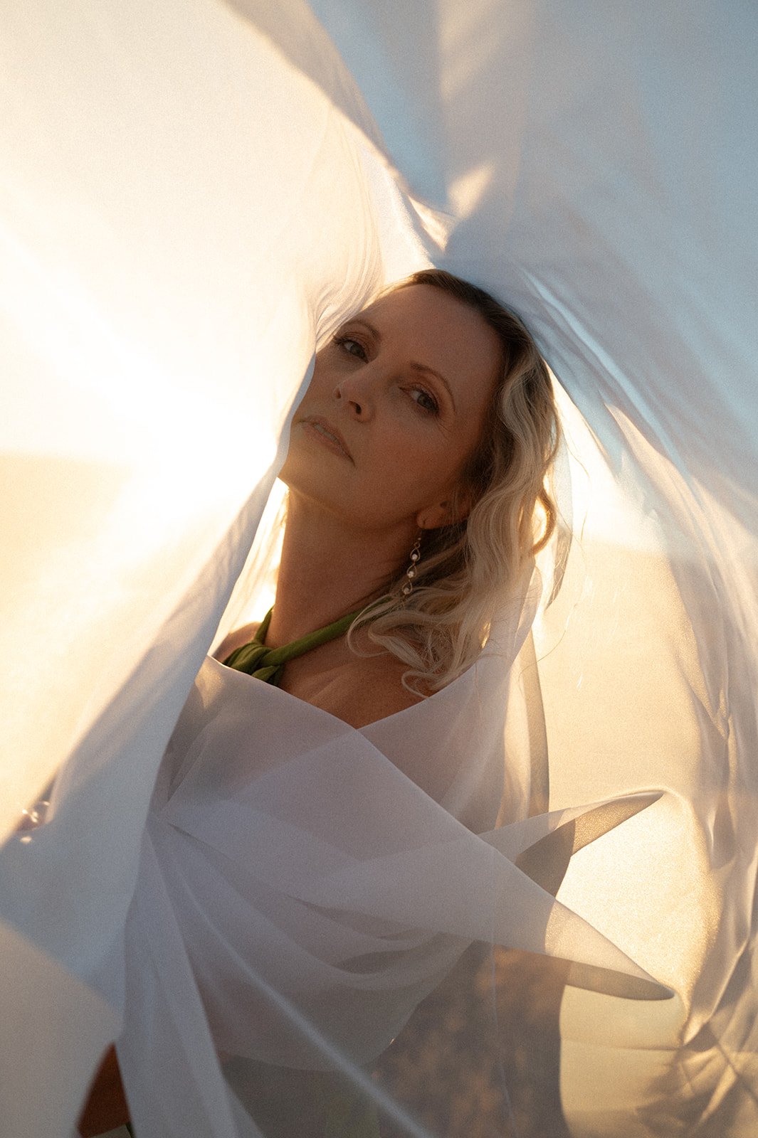 A woman with blonde hair and earrings shown through white fabric, possibly taking a selfie in sunlight.
