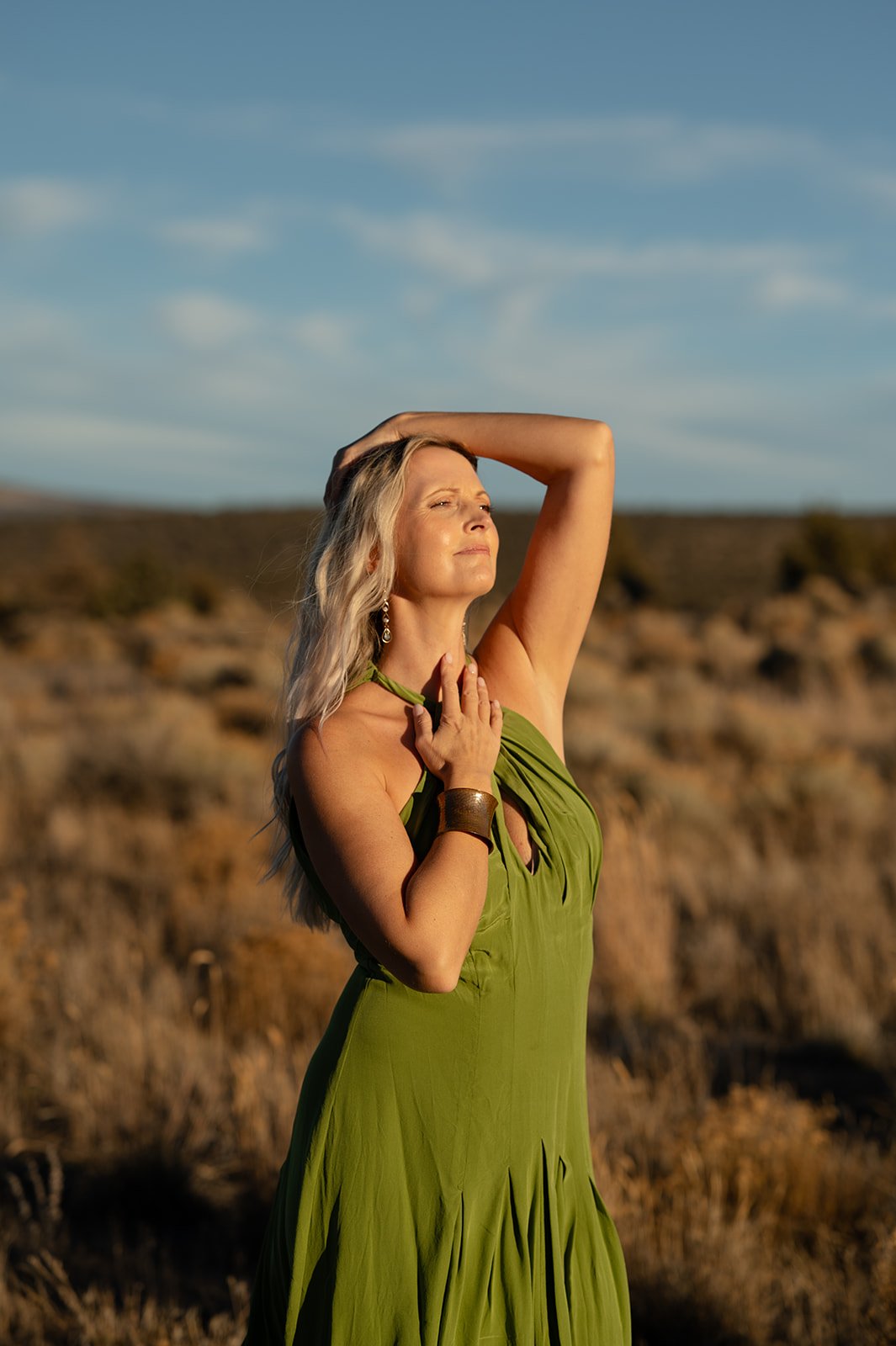 A woman with blonde hair wearing a green dress, with her eyes closed and one arm raised, standing outdoors in a dry, grassy landscape under a blue sky.