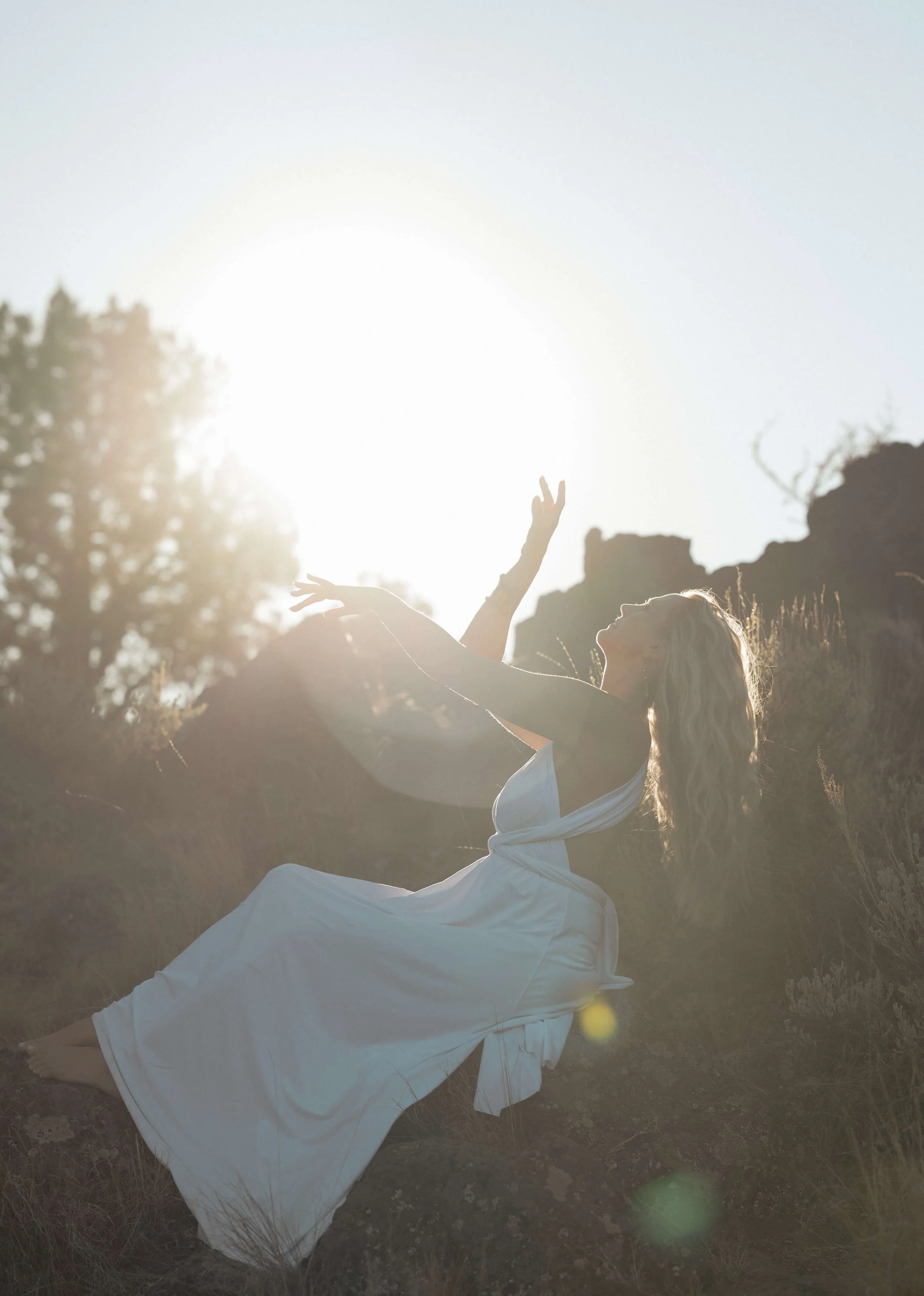 A woman in a white dress sitting outdoors at sunset, reaching towards the sun with her arms extended and head tilted back.