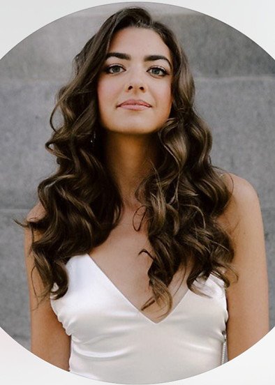 A young woman with long, wavy dark hair and light skin, wearing a white satin top, posing in front of a gray background.
