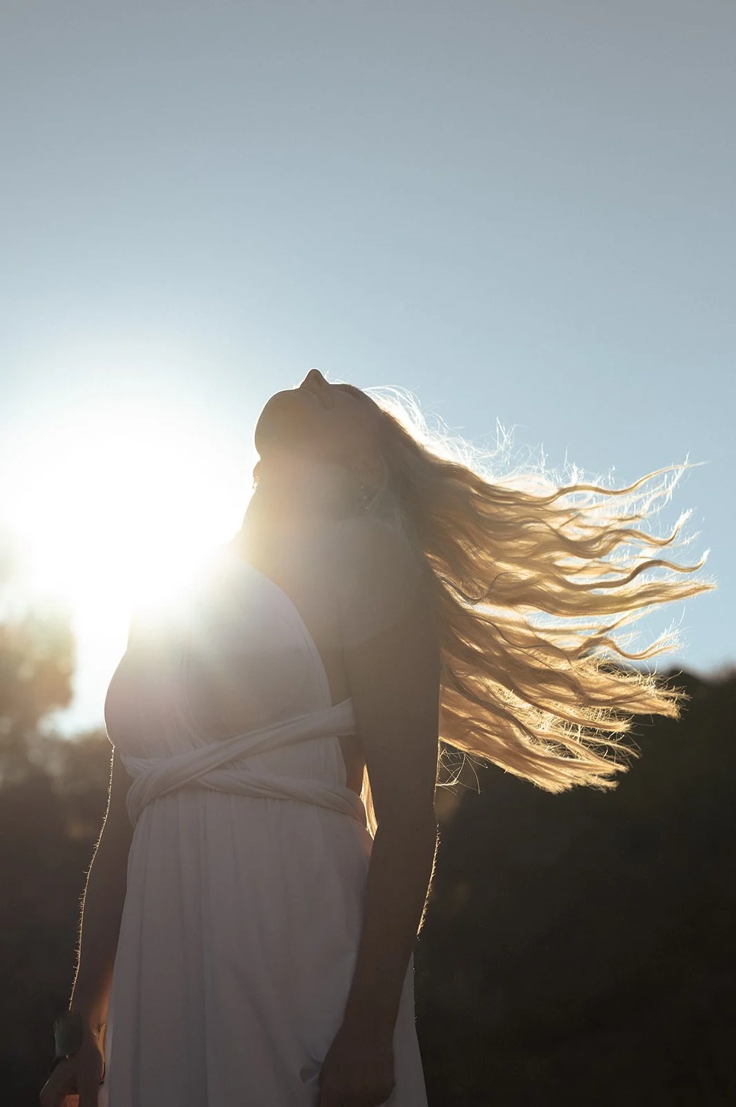 Woman with long, flowing hair standing outdoors with the sun behind her, creating a backlit silhouette.