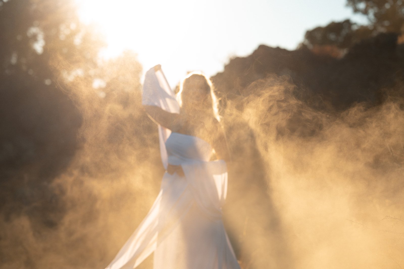 A woman in a flowing white dress and veil walking through a cloud of dust or mist, backlit by the sun, creating a warm, glowing atmosphere.
