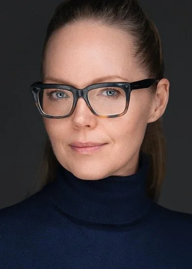 A woman with brown hair in a ponytail, wearing glasses and a dark turtleneck, looking at the camera against a dark background.