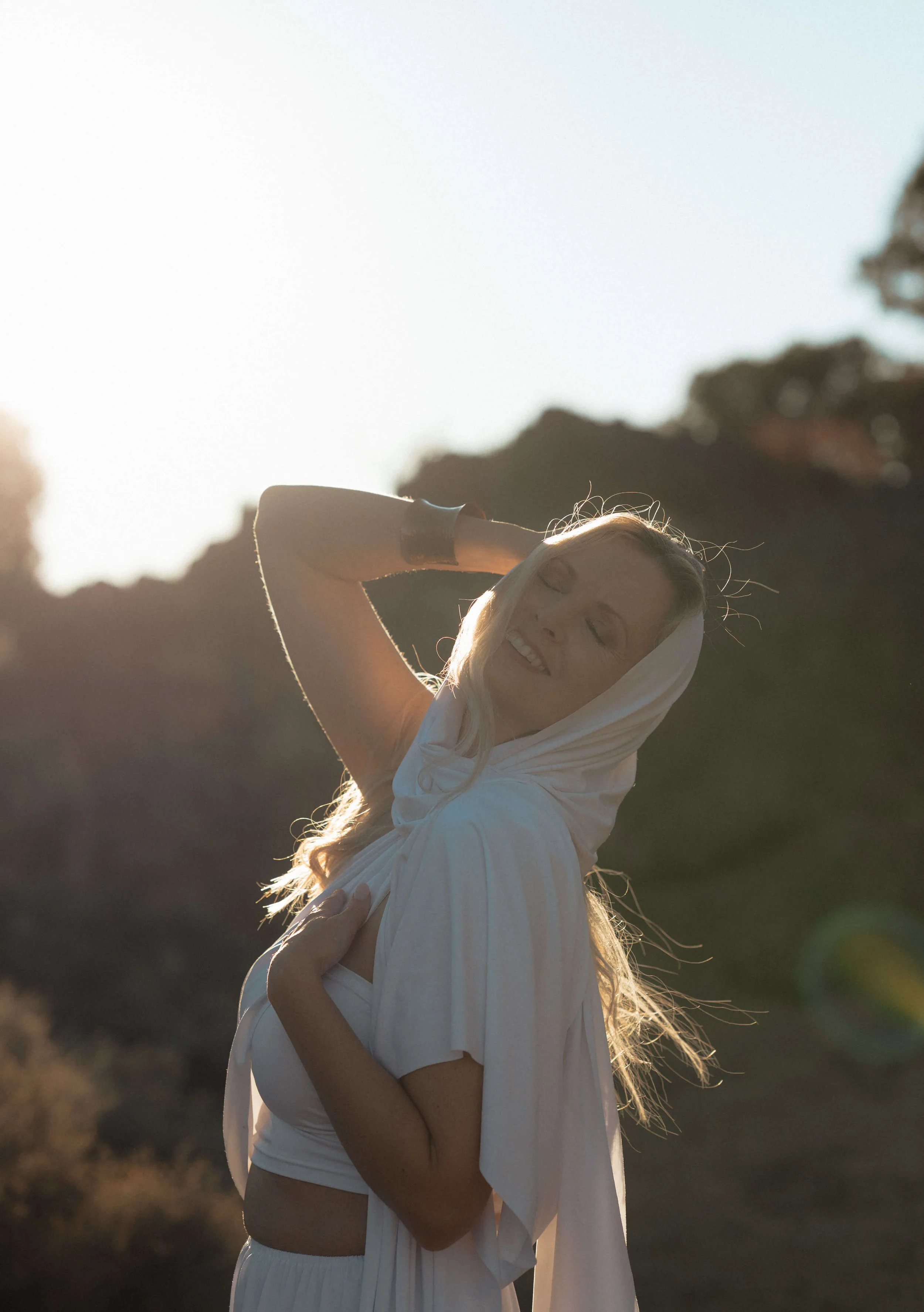 Woman smiling with eyes closed, wearing light-colored clothing and a head covering, standing outdoors during sunset.