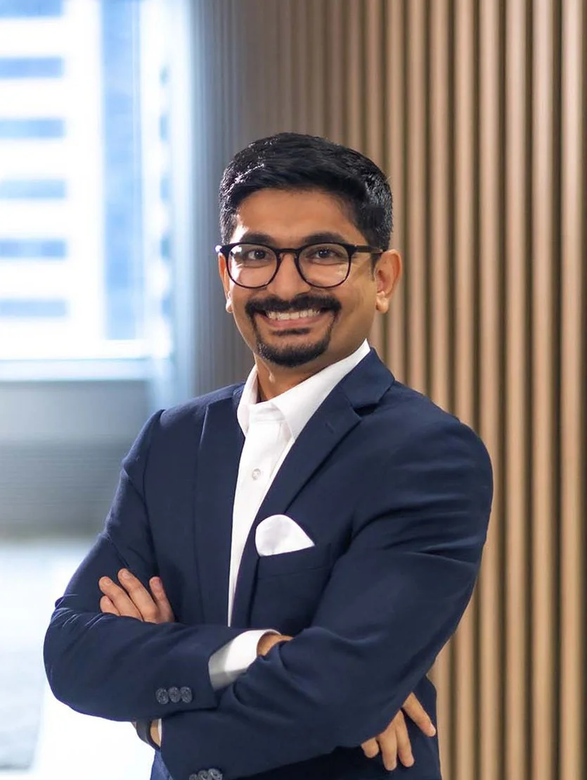Akhil France, financial adviser in a dark blue suit with a white shirt and pocket square, wearing glasses, smiling with arms crossed in an office setting with wooden vertical slats and a window in the background.