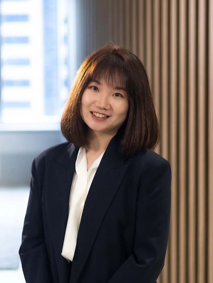 Lynn Lin, financial adviser with shoulder-length brown hair, wearing a black blazer and white blouse, smiling in an indoor setting with a window and wooden paneling in the background.