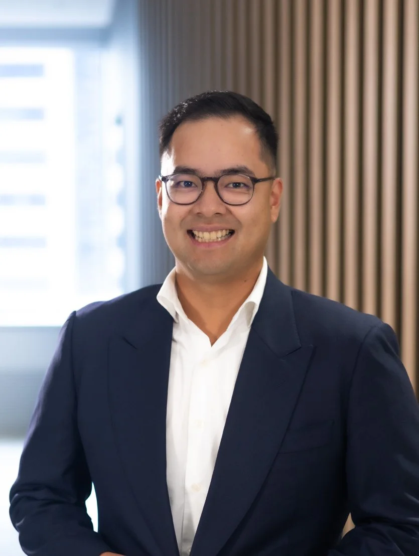 Angelo Yao, finance broker in a navy suit and white shirt, wearing glasses, standing in an indoor office with wooden paneling and windows.