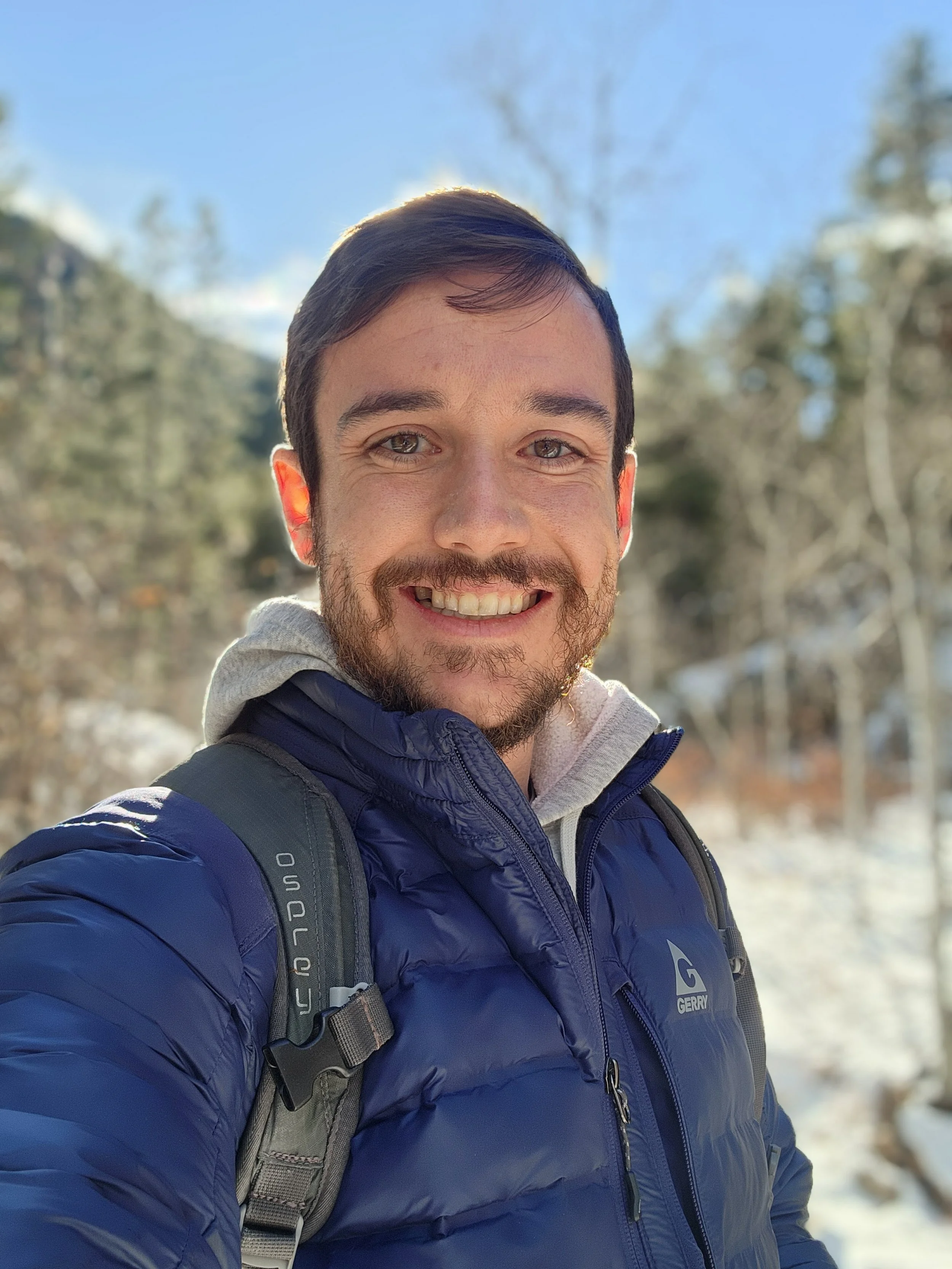 A smiling man with a beard taking a selfie outdoors in a snowy, wooded area during daytime.