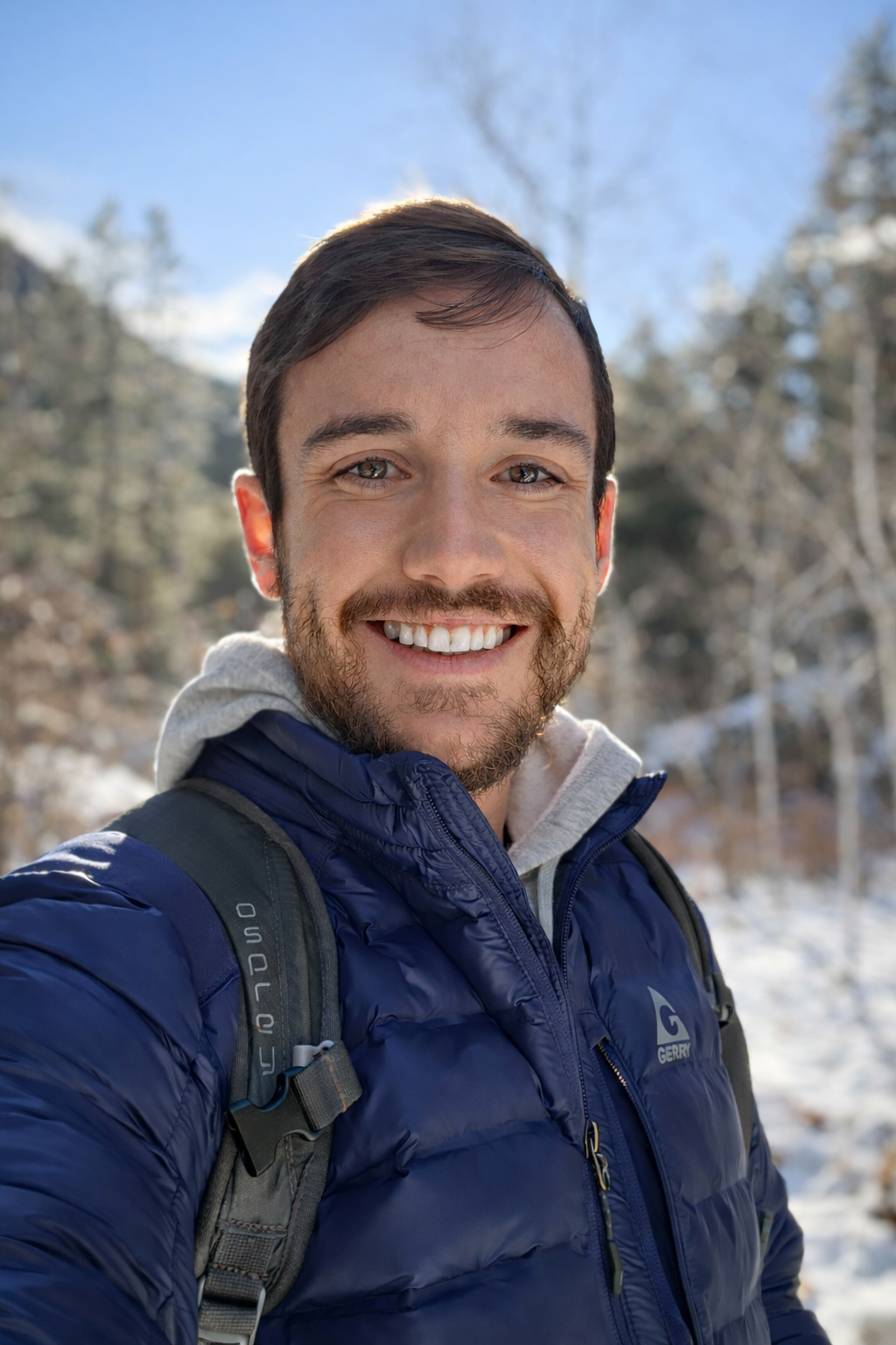 A smiling man with light skin and dark hair outdoors in a snowy forest, wearing a blue jacket and carrying a backpack.