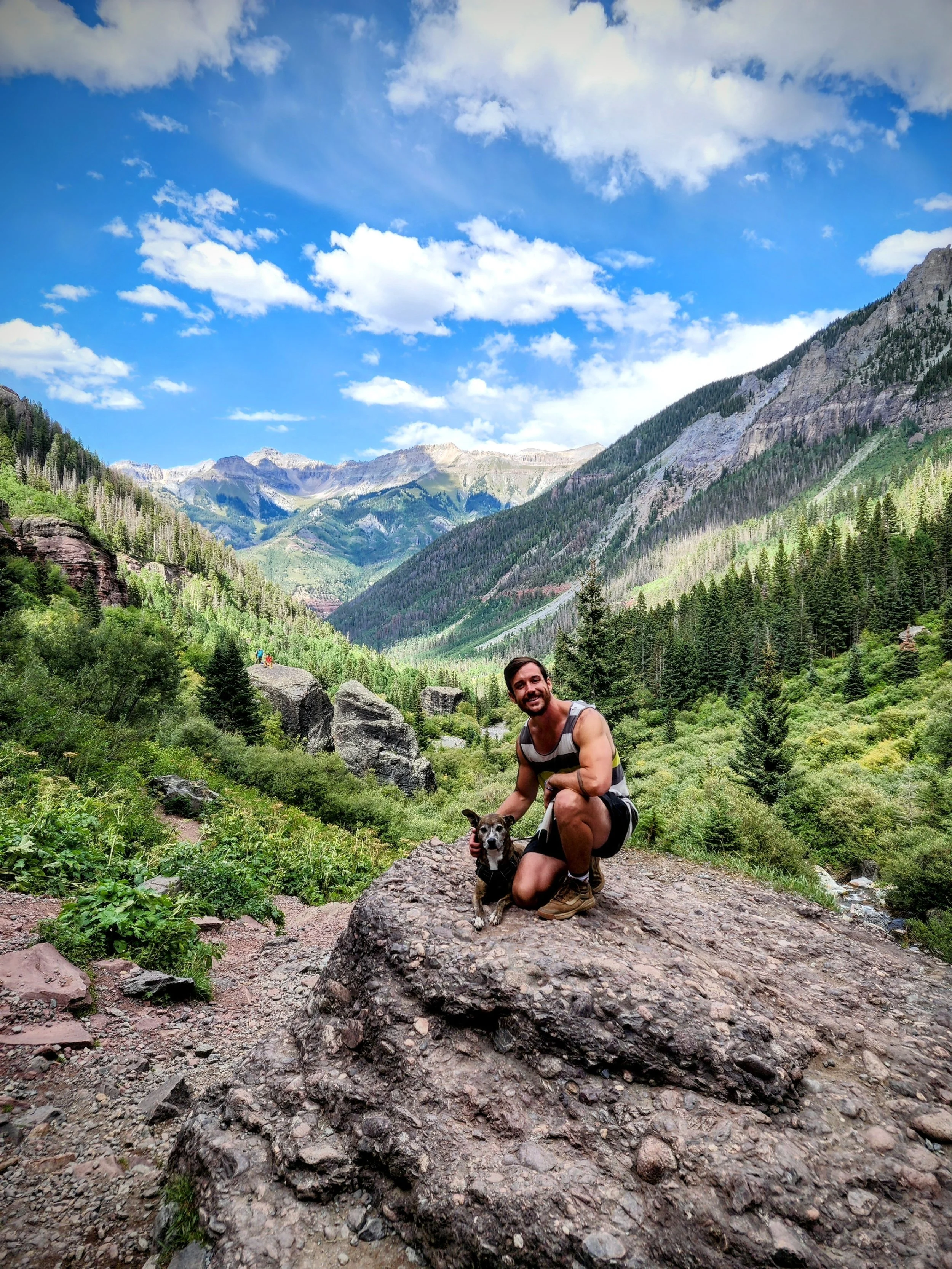 A man kneeling on a large rock in a lush green valley with mountains and a blue sky with clouds in the background, holding a small dog.