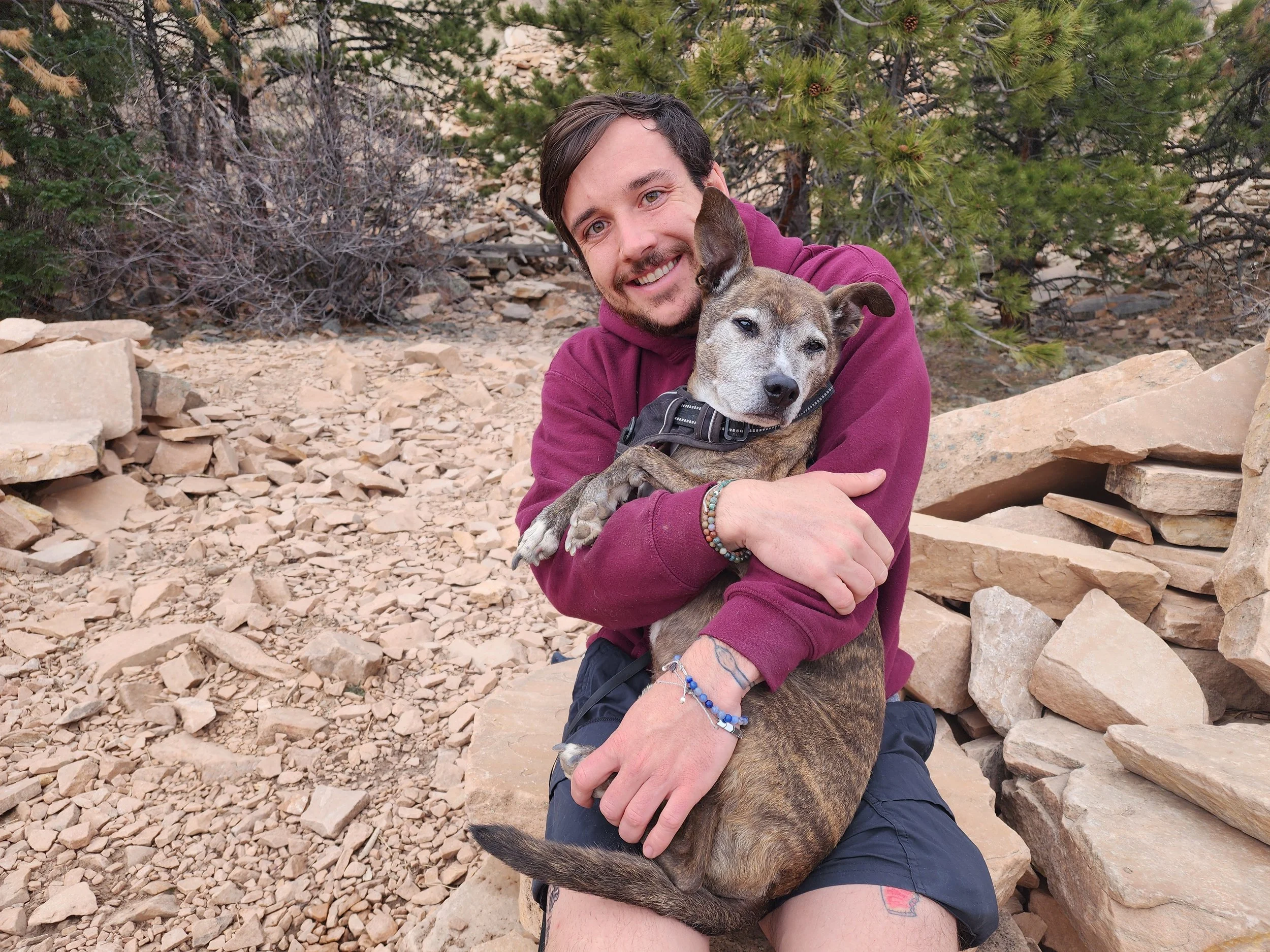 A man with a beard and mustache, wearing a maroon hoodie and black shorts, is sitting on a rocky trail, smiling and hugging a brindle dog with a black collar. The background features green pine trees and rocky terrain.
