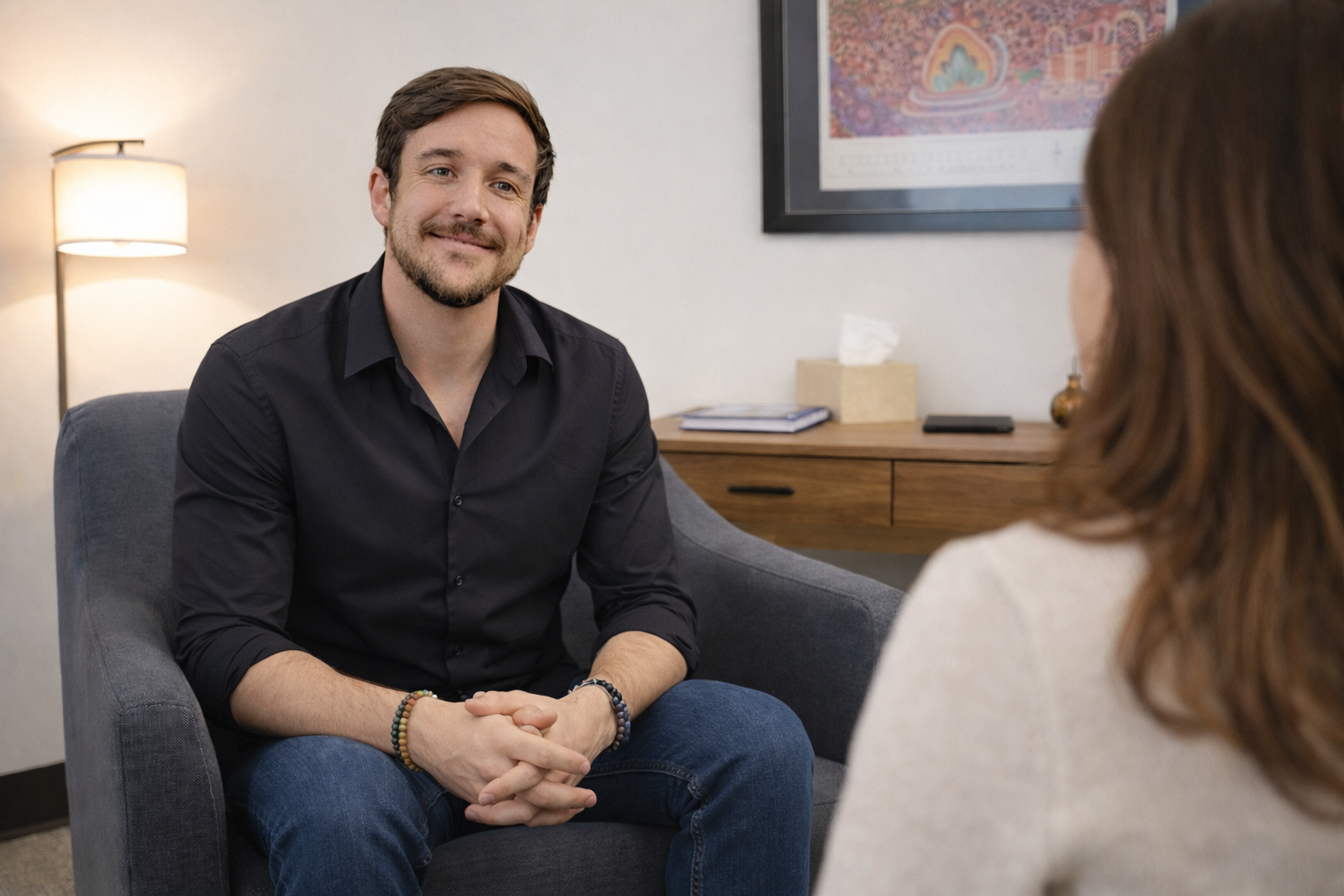 A man with dark hair, beard, and mustache, wearing a black shirt and blue jeans, sitting in a gray armchair, smiling and talking to a woman with long brown hair, wearing a white top, in an office or therapy room with a framed picture on the wall, a wooden desk with a tissue box, a notebook, and a phone, and a standing lamp.