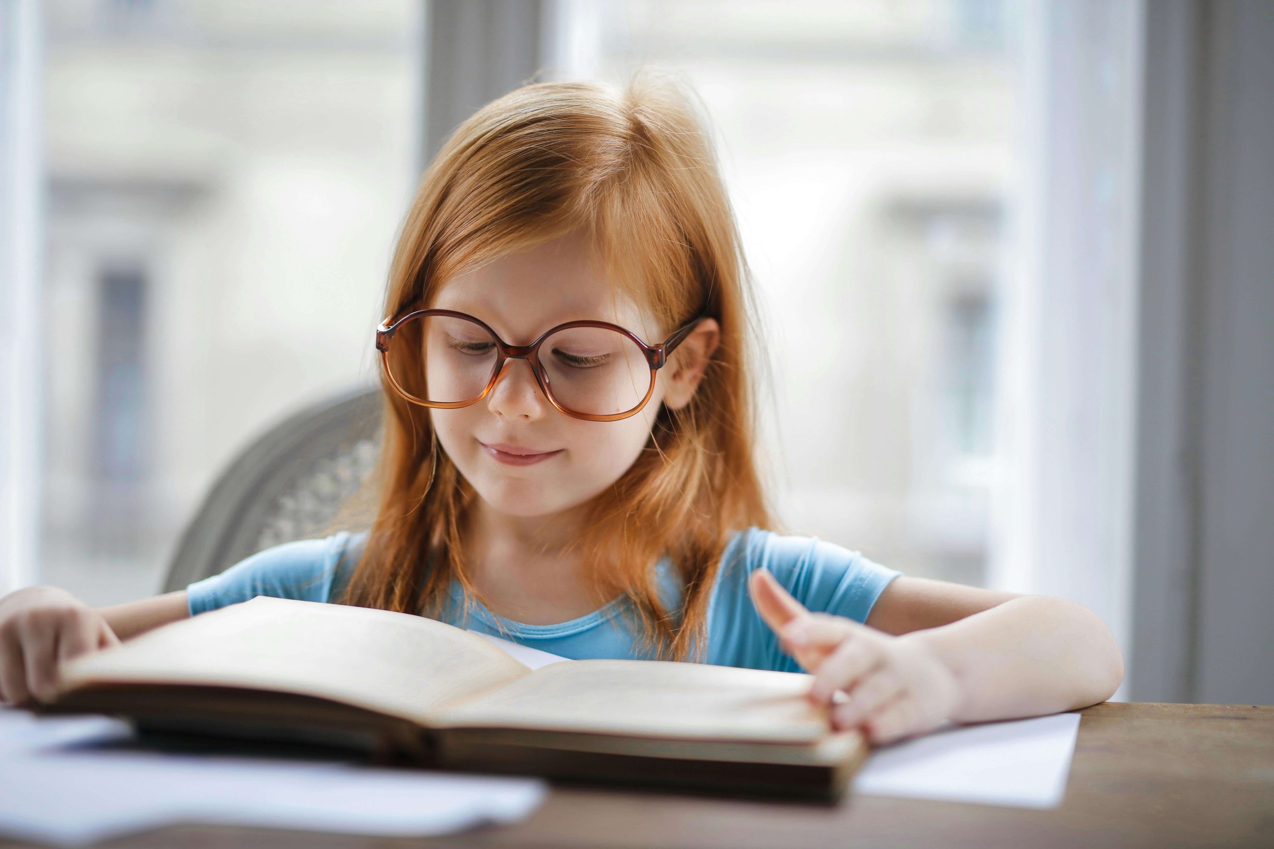 Young girl with red hair and glasses reading a book at a desk near a window.