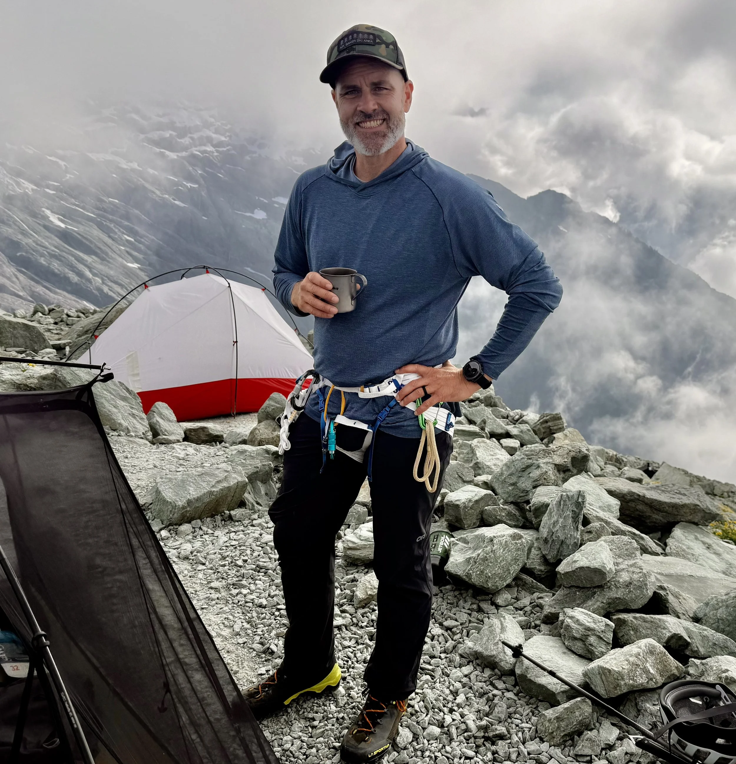 A man stands on a rocky mountain summit holding a cup, wearing outdoor gear and climbing harness, with a red and white tent behind him amidst clouds and mountain peaks.