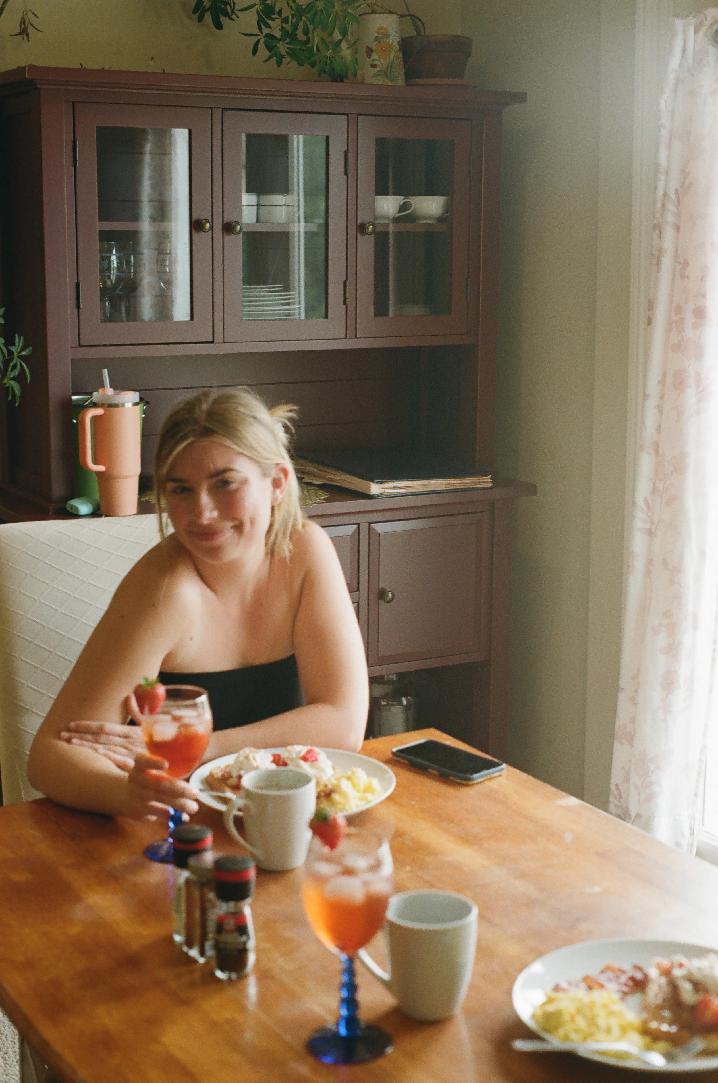 A woman with blonde hair smiling at a dining table with plates of breakfast foods, cups, and glasses of pink beverage, in a cozy kitchen with a wooden cabinet in the background.