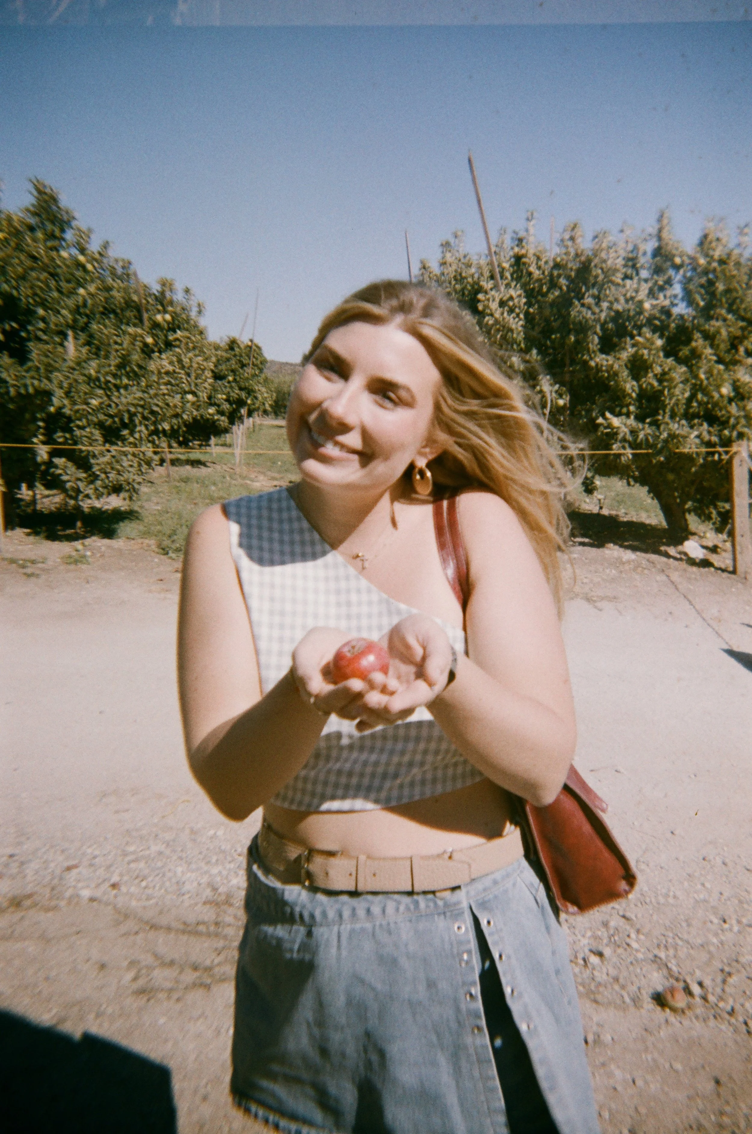 A smiling woman with long blonde hair holding apples outdoors on a sunny day, with greenery and a clear blue sky in the background.