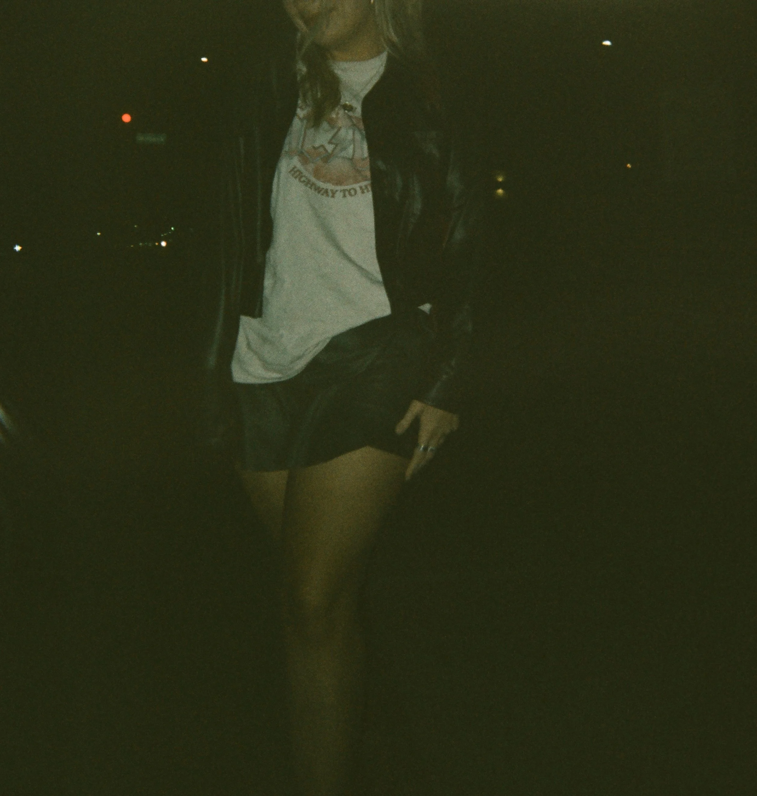 Person wearing a black leather jacket, graphic t-shirt, black shorts, and black tights standing outdoors at night, holding a phone.
