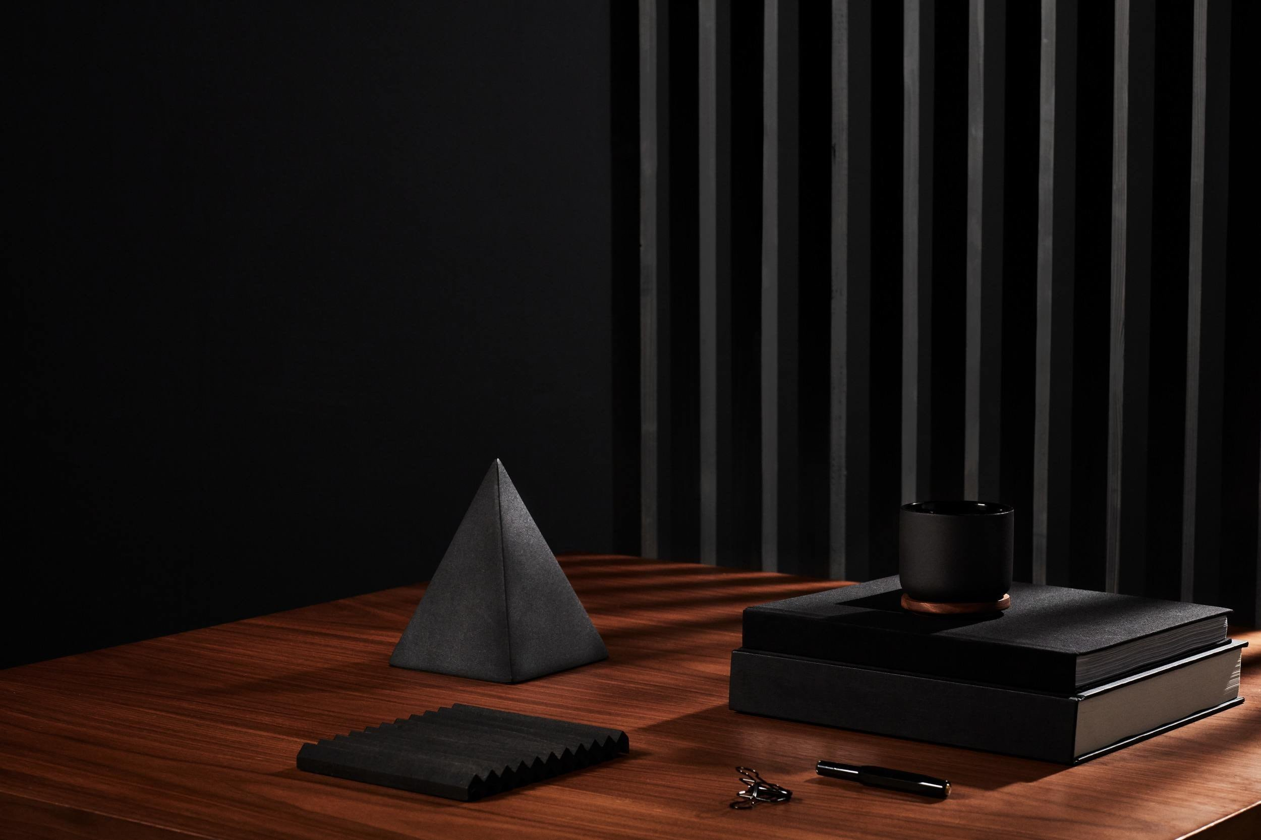 Surface en bois avec un objet pyramidal gris, un carnet noir, un mug noir, une pile de deux autres carnets, des ciseaux, un stylo noir, et un échantillon de tissu noir, tous disposés contre un fond noir avec un panneau en lamelles verticales.