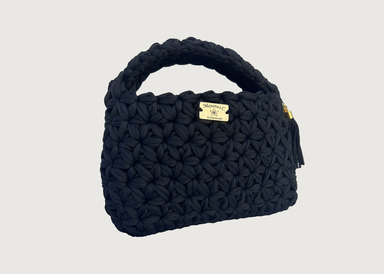 Sac à main noir en tissu tressé avec une étiquette en métal doré portant le nom 'Mayana's C.' et la mention 'HANDMADE', doté d'une fermeture éclair avec une tirette en or.