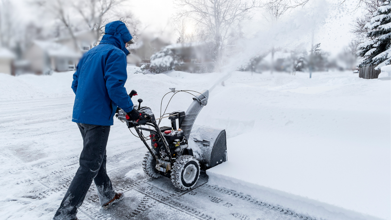 A person wearing a blue winter coat and black pants, using a snow blower on a snowy residential street during daylight.