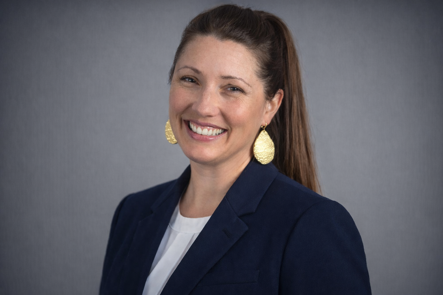 A professional woman with brown hair smiling, wearing a navy blazer, white blouse, and large gold earrings, against a gray background.