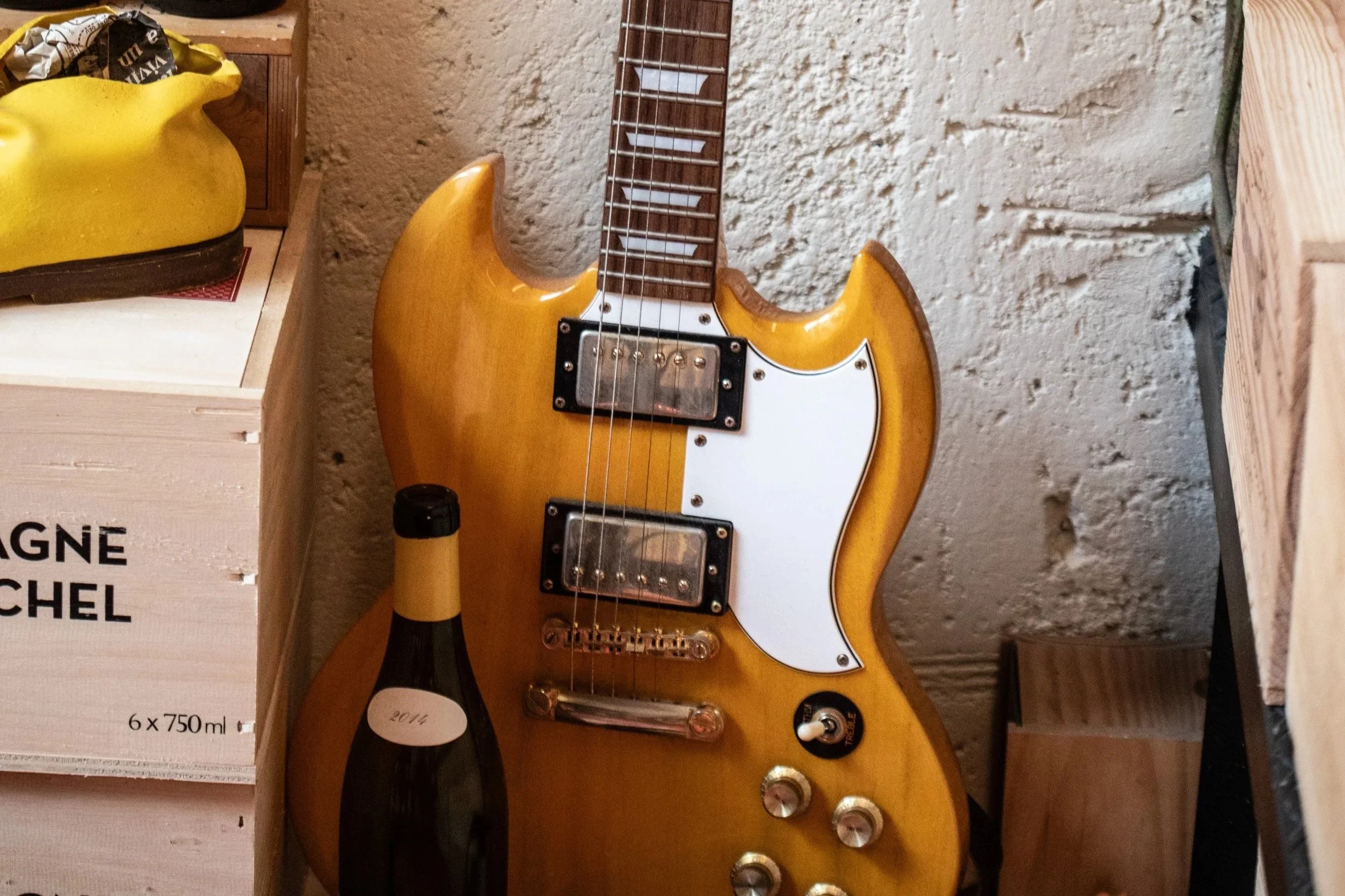 Chitarra elettrica di colore giallo scuro con manico di legno, due pickup, effetti e manopole, posizionata vicino a una bottiglia di vino e a casse di vino su una parete di pietra.