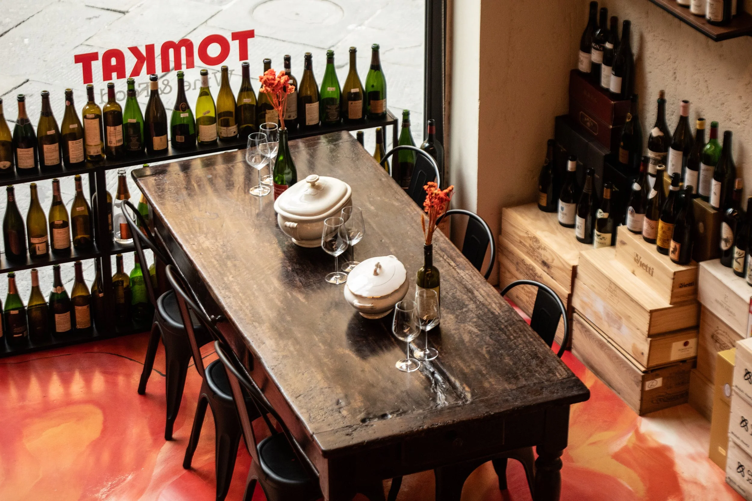 Interno di una sala con tavolo in legno, bicchieri di vino, vasi con fiori e scaffali di bottiglie di vino.