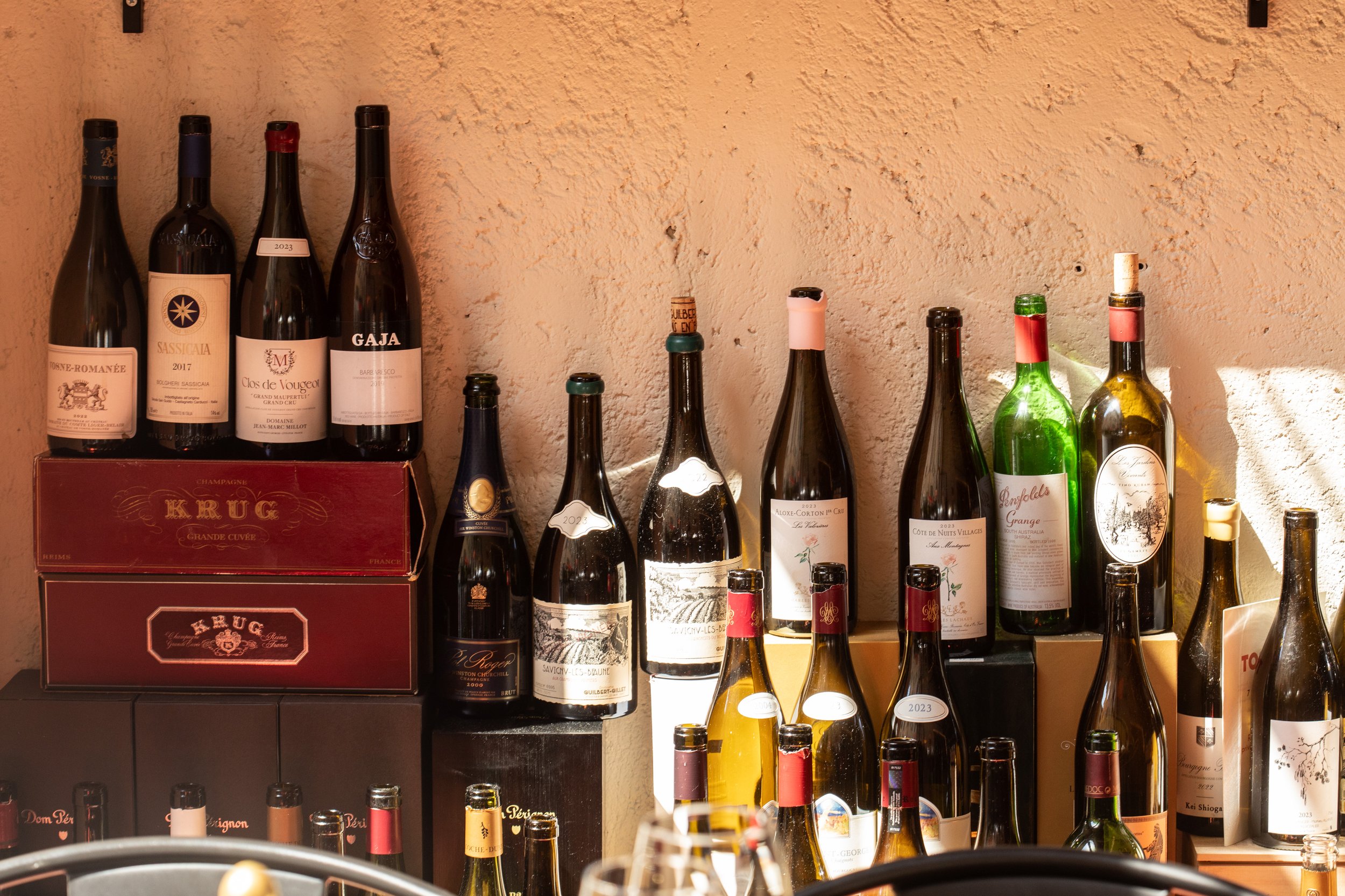 Bottiglie di vino su scaffali, con alcuni in una scatola rossa sotto. Parete di muro color pesca sullo sfondo.