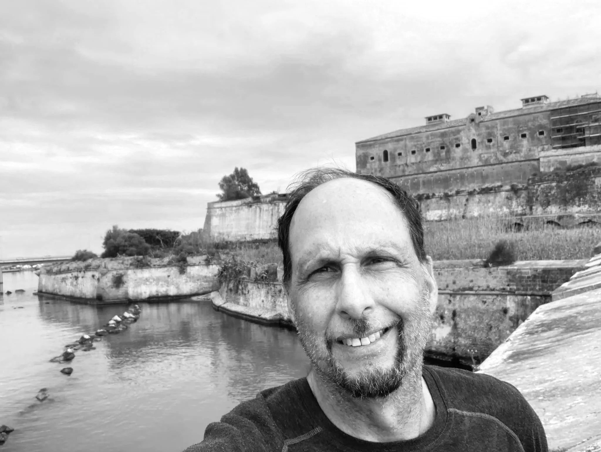 A smiling man, Gregg Orifici, taking a selfie near a historic fort by the water, with a cloudy sky overhead.