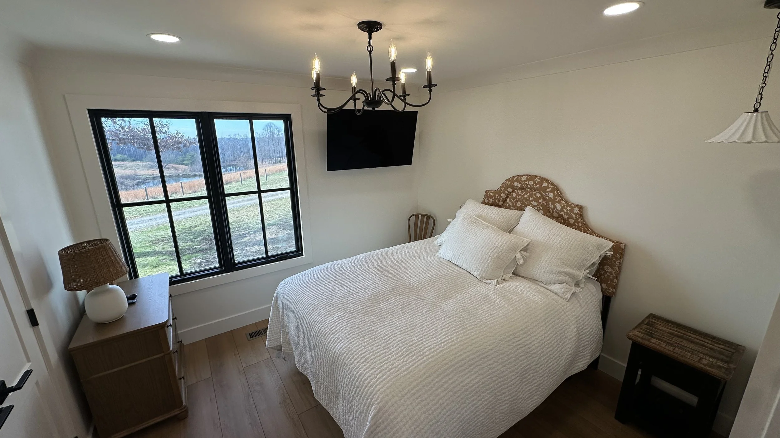 A cozy bedroom with a large bed covered in a white quilt and matching pillows. There is a wooden nightstand with a white lamp on the left side, a small wooden chair beside the bed, and a window with black framing showing an outdoor scene of grass, tr