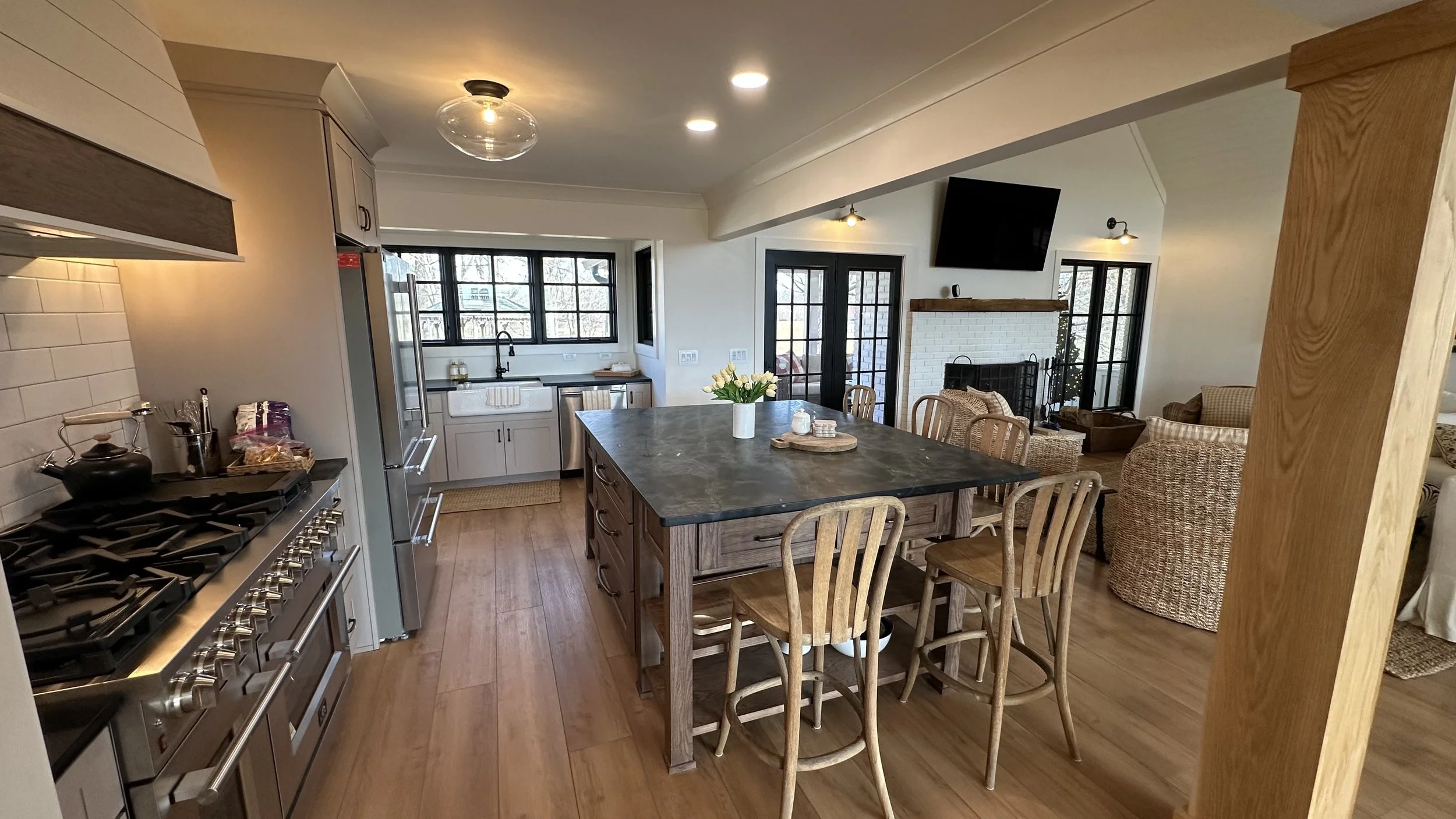 Open kitchen and living room with wooden flooring, dining table with chairs, kitchen with white cabinets, stainless steel appliances, and a living area with wicker and upholstered chairs, a fireplace, and a wall-mounted TV.