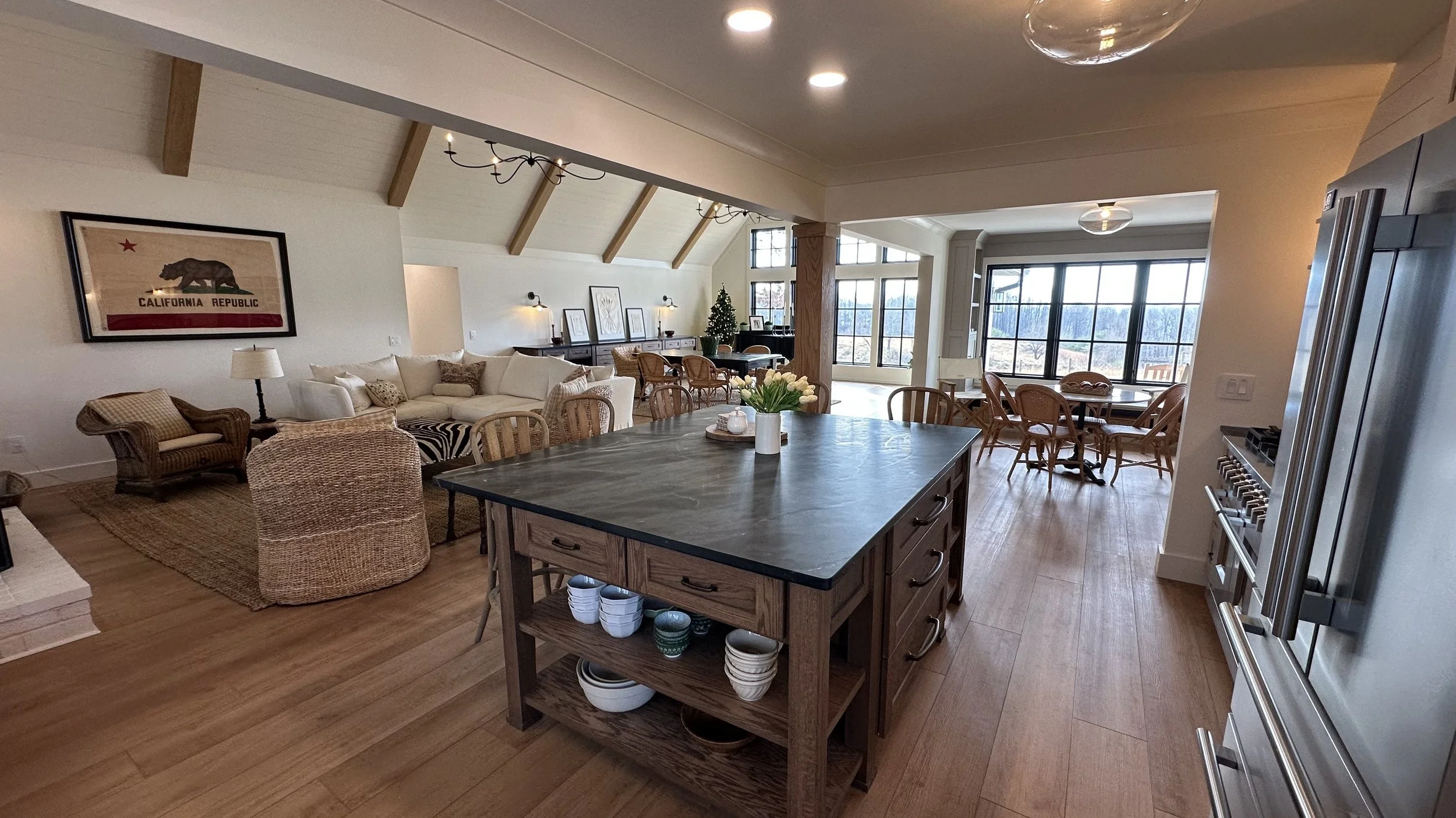 Open-concept living space with kitchen island, dining area, and sitting area with white sofa, armchair, and artwork. Large windows with a view outside.