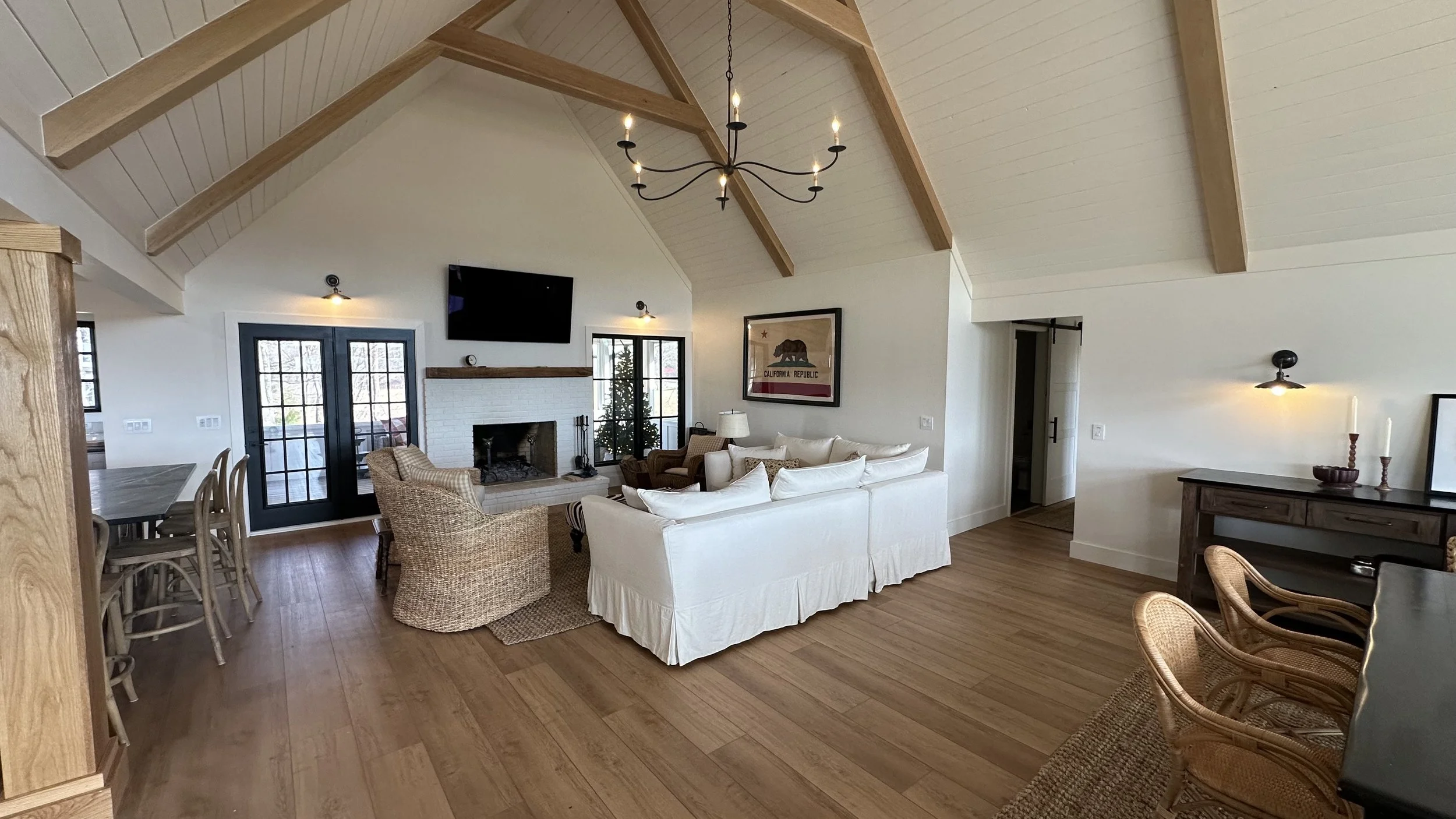 Spacious living room with vaulted ceiling, wooden beams, chandelier, fireplace, flat-screen TV, and seating area with white sofa, armchairs, and wooden chairs.