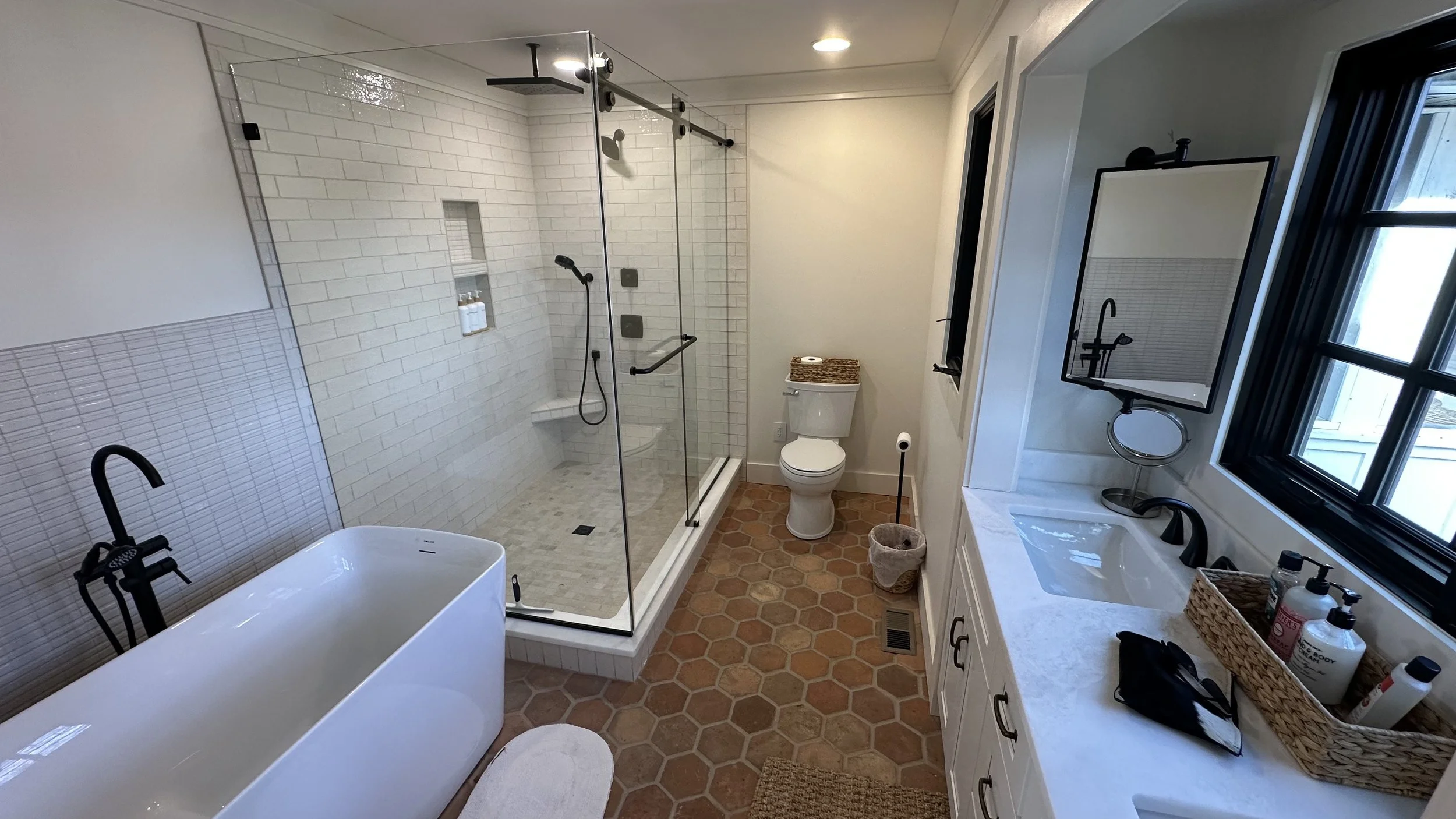Bathroom with a freestanding bathtub, a glass-enclosed shower, a toilet, a sink with a mirror above it, a window, and various toiletries on the countertop.