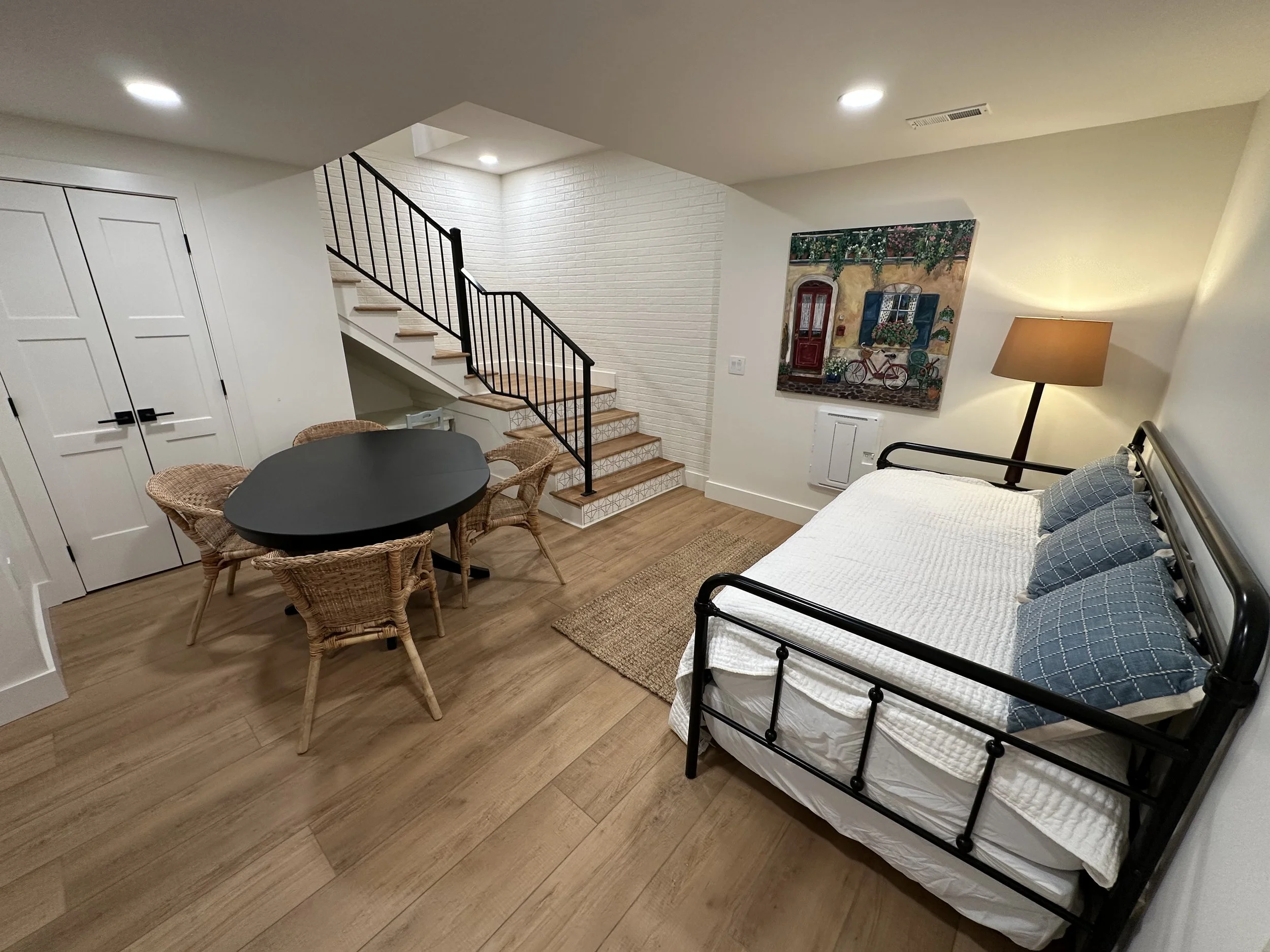 Living space with a black metal bed frame, a nightstand with a lamp, a painting on the wall, a wicker table with chairs, and a staircase with black railing.