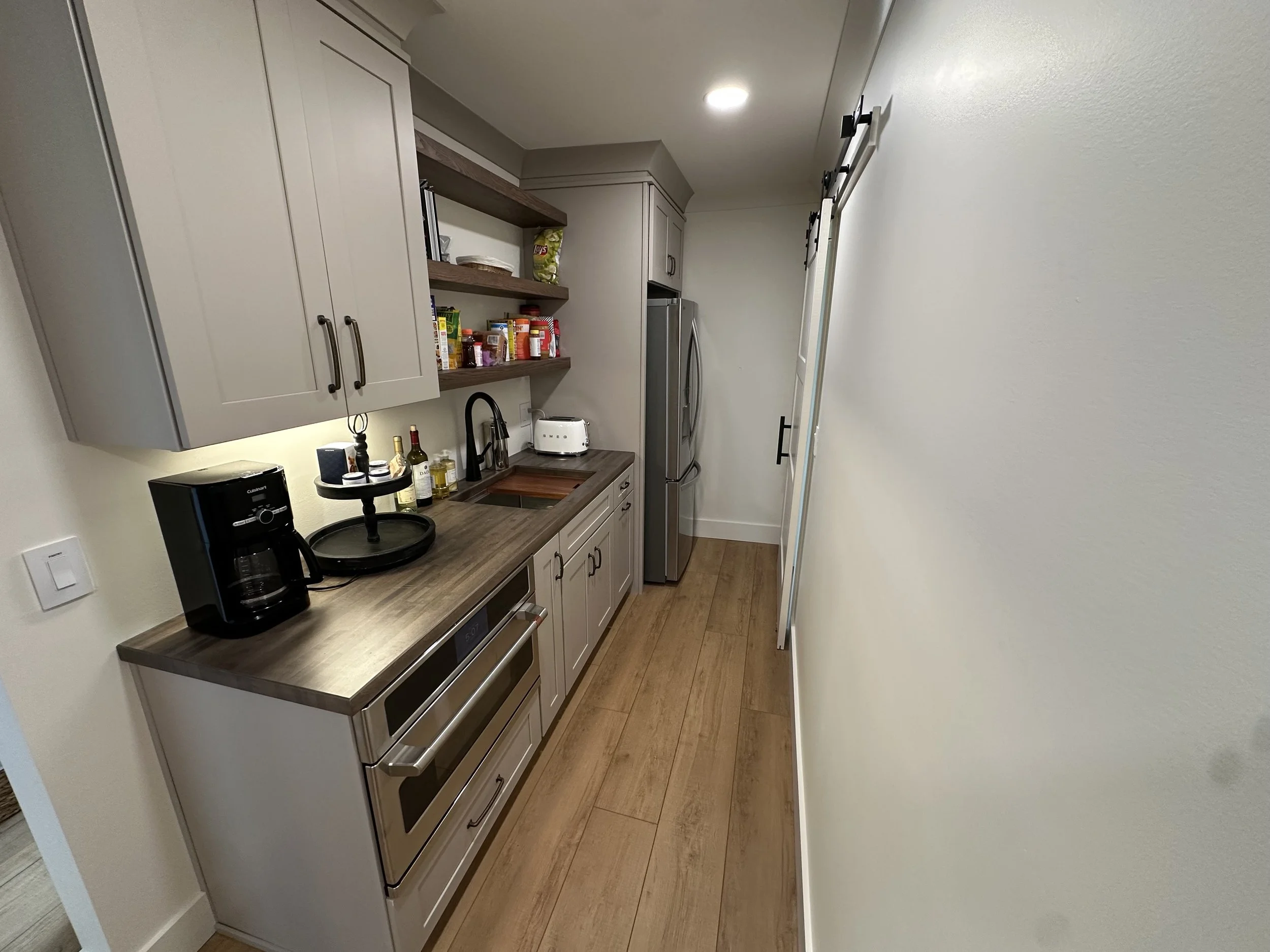 Narrow kitchen with gray cabinets, wooden countertop, open shelves holding pantry items, stainless steel oven and refrigerator, coffee maker, toaster, and a black tiered serving tray on the counter.